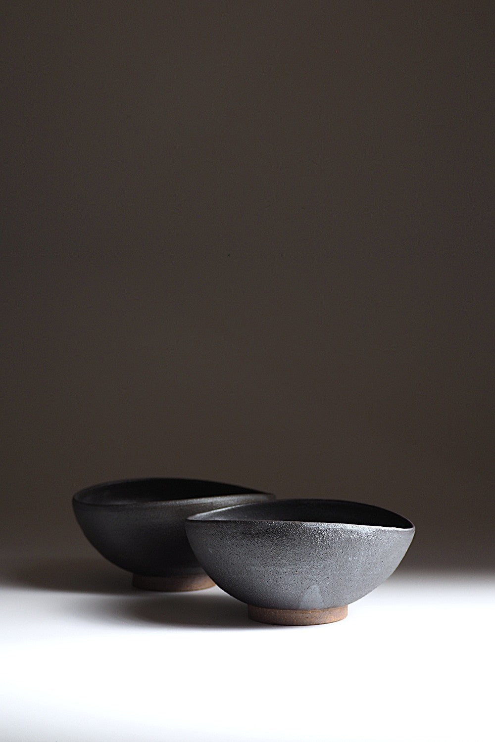 Two black handcrafted Japanese ceramic bowls on a white surface with a dark background, Tsukushi