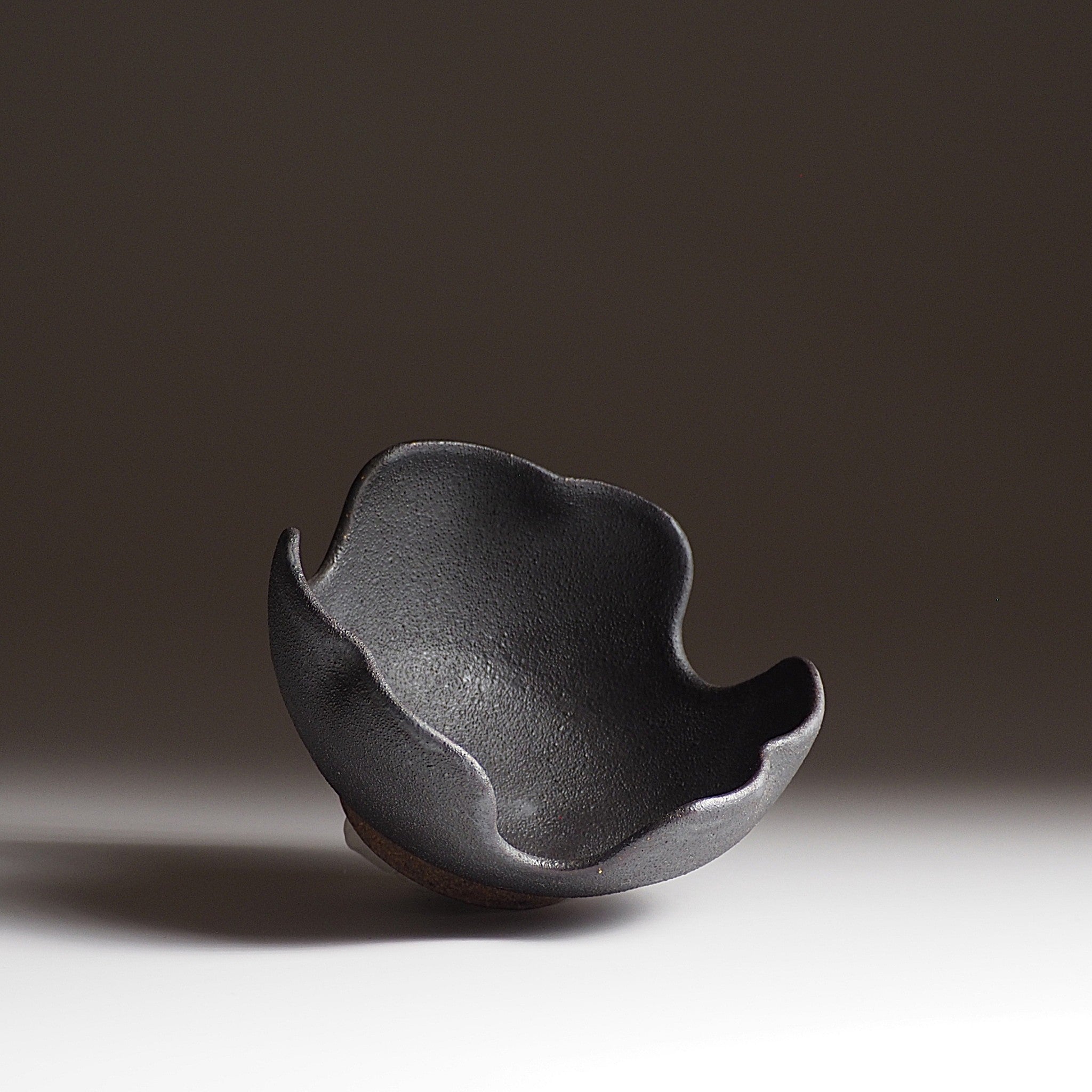 Side view of a black Japanese pottery bowl shaped like a flower. Tsukushi