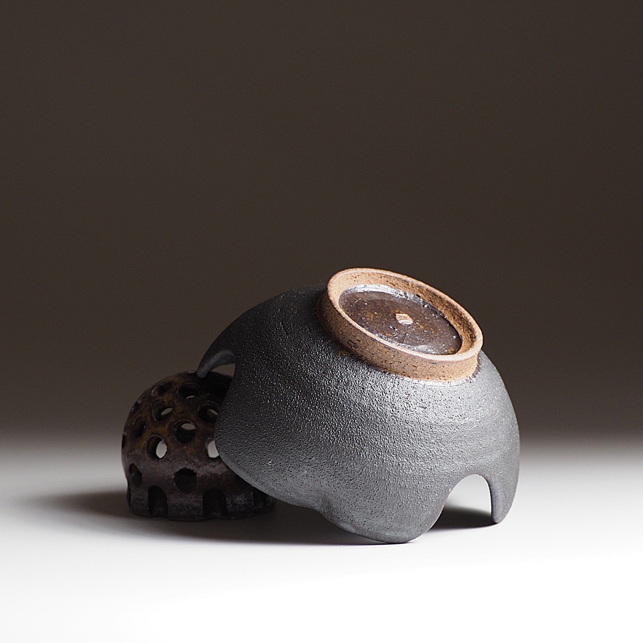 Bottom view of an unglazed pottery bowl lying diagonally on a ceramic flower frog. Tsukushi.