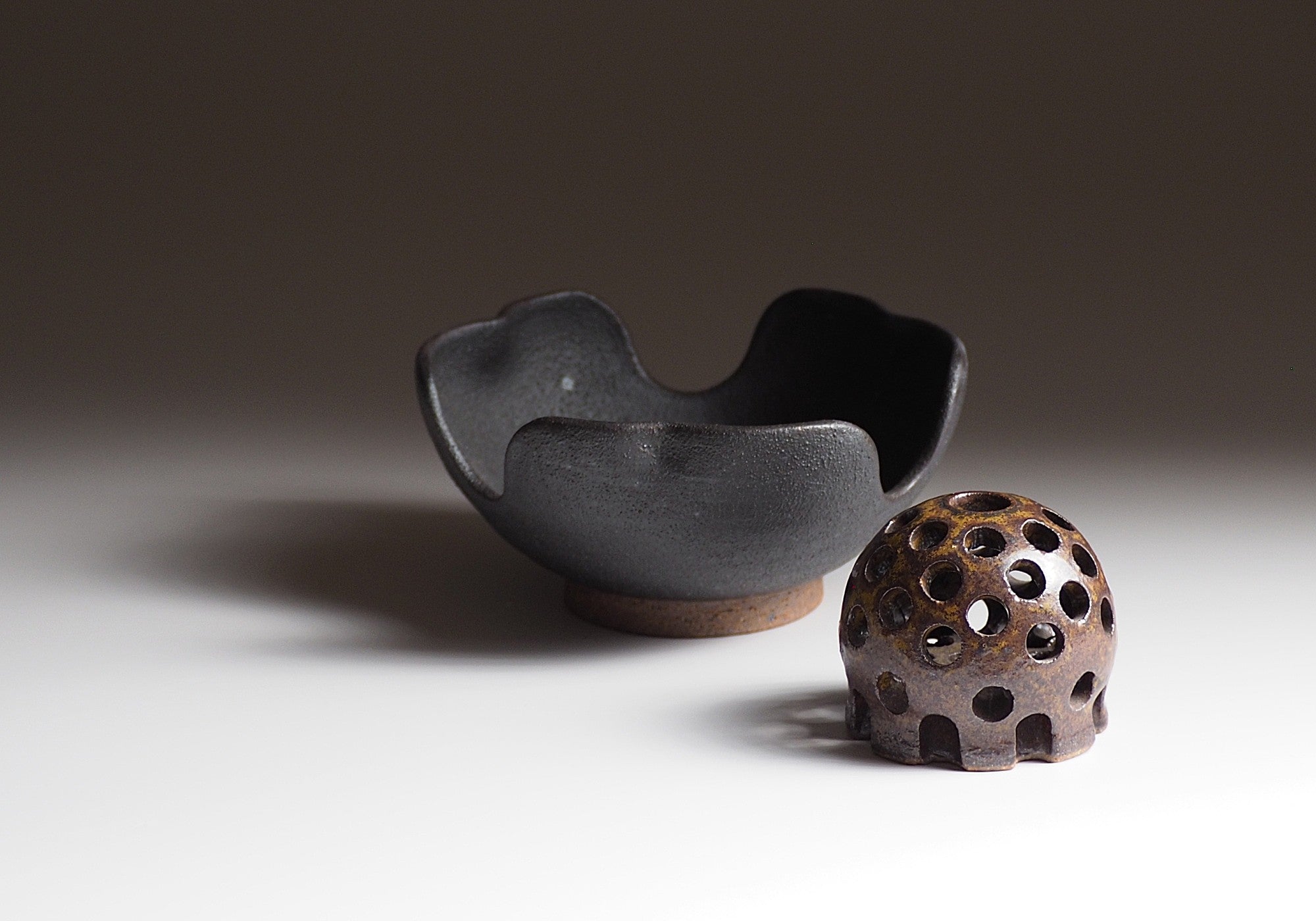 Two Japanese ceramic items, one flower black bowl and one flower frog, on a neutral background.