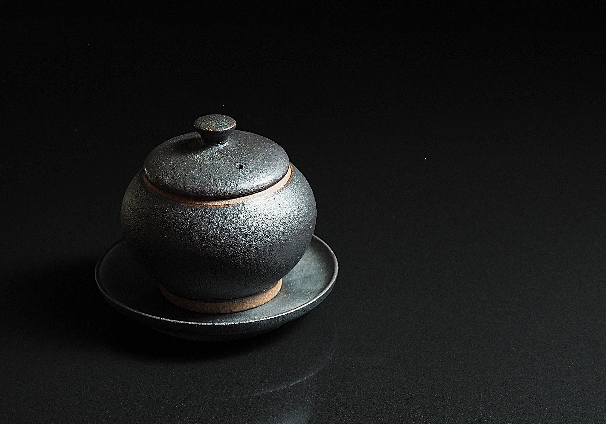 Bird’s-eye view of a black chawanmushi bowl made of unglazed pottery, set against a faded background. Tsukushi.