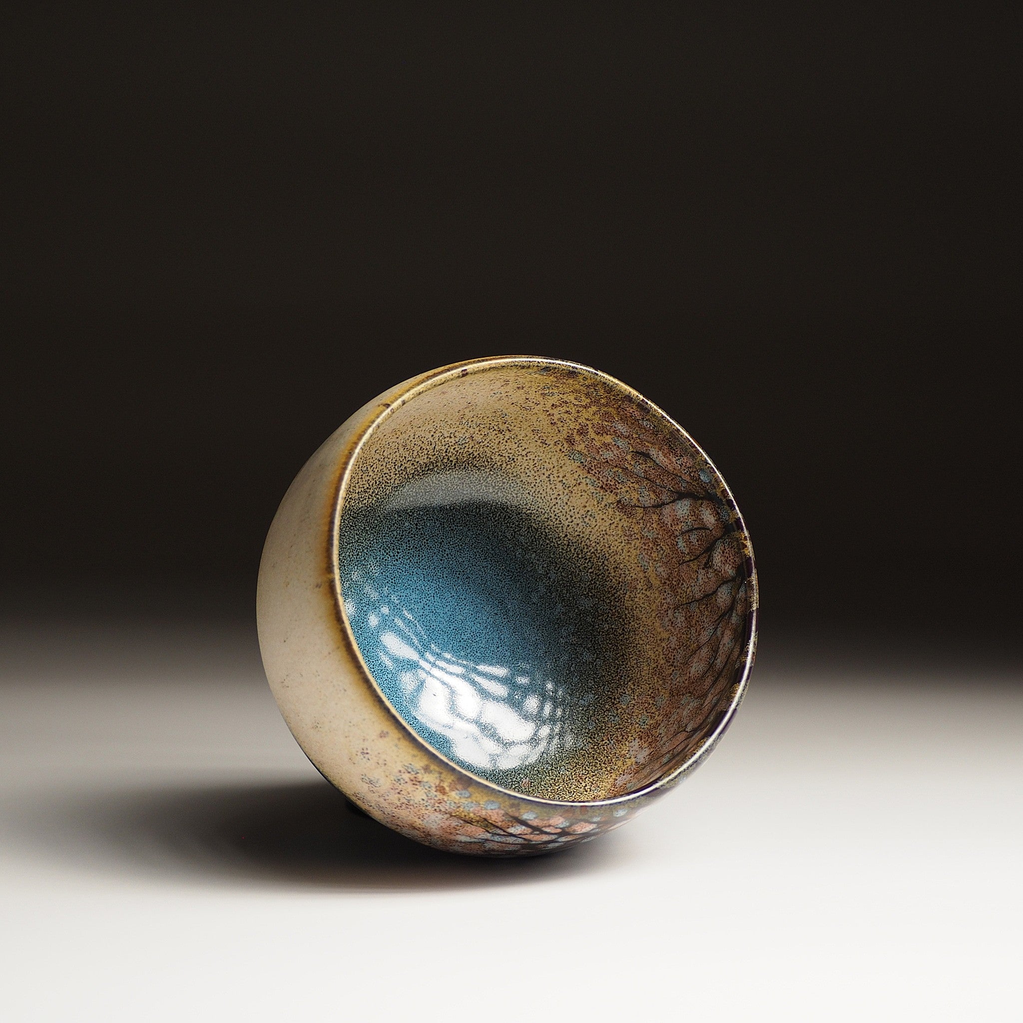 A top view of a Japanese ceramic matcha bowl designed with cherry blossoms and a tranquil lake against a black background. Tsukushi