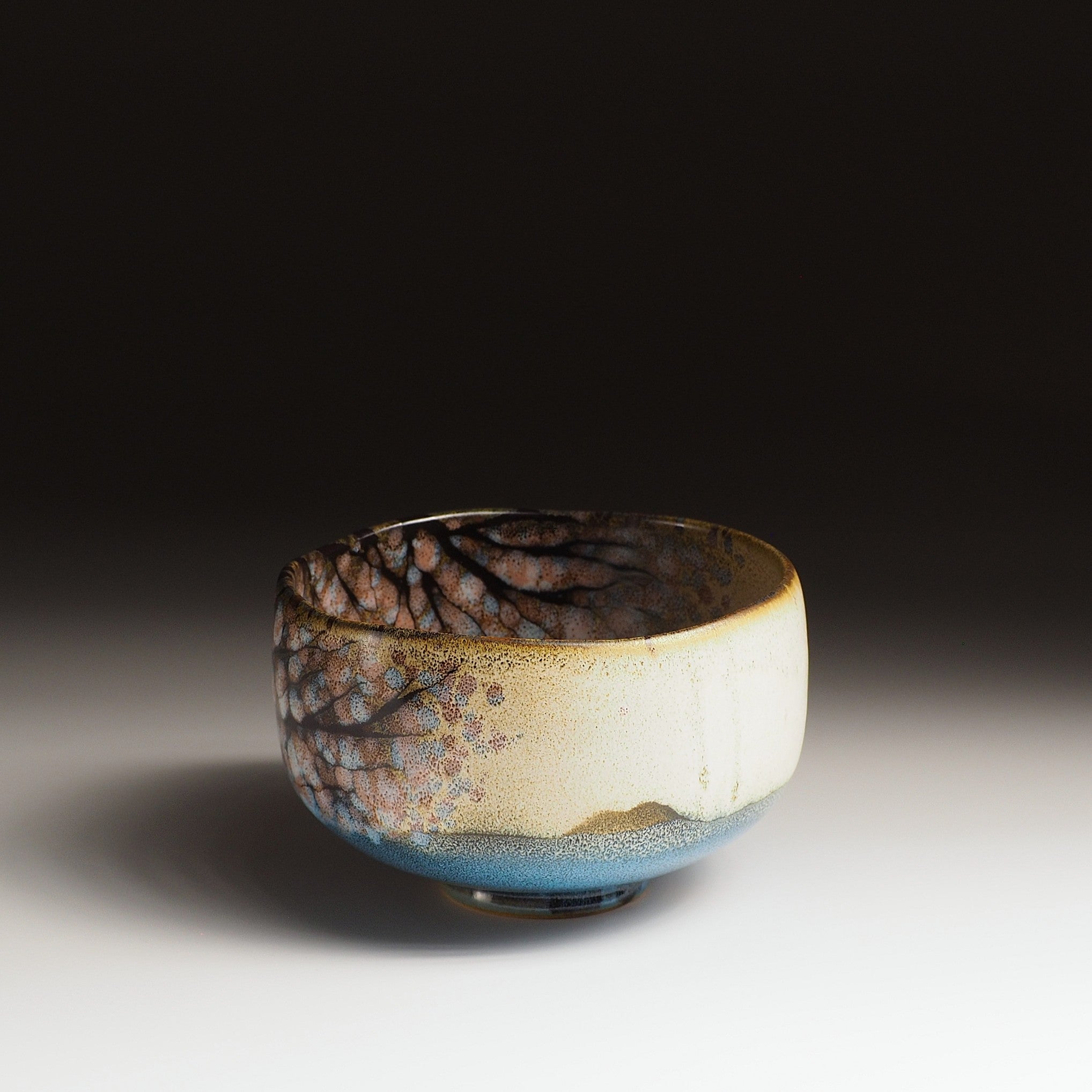 A side view of a Japanese ceramic matcha bowl adorned with cherry blossoms and a lake against a black background. Tsukushi