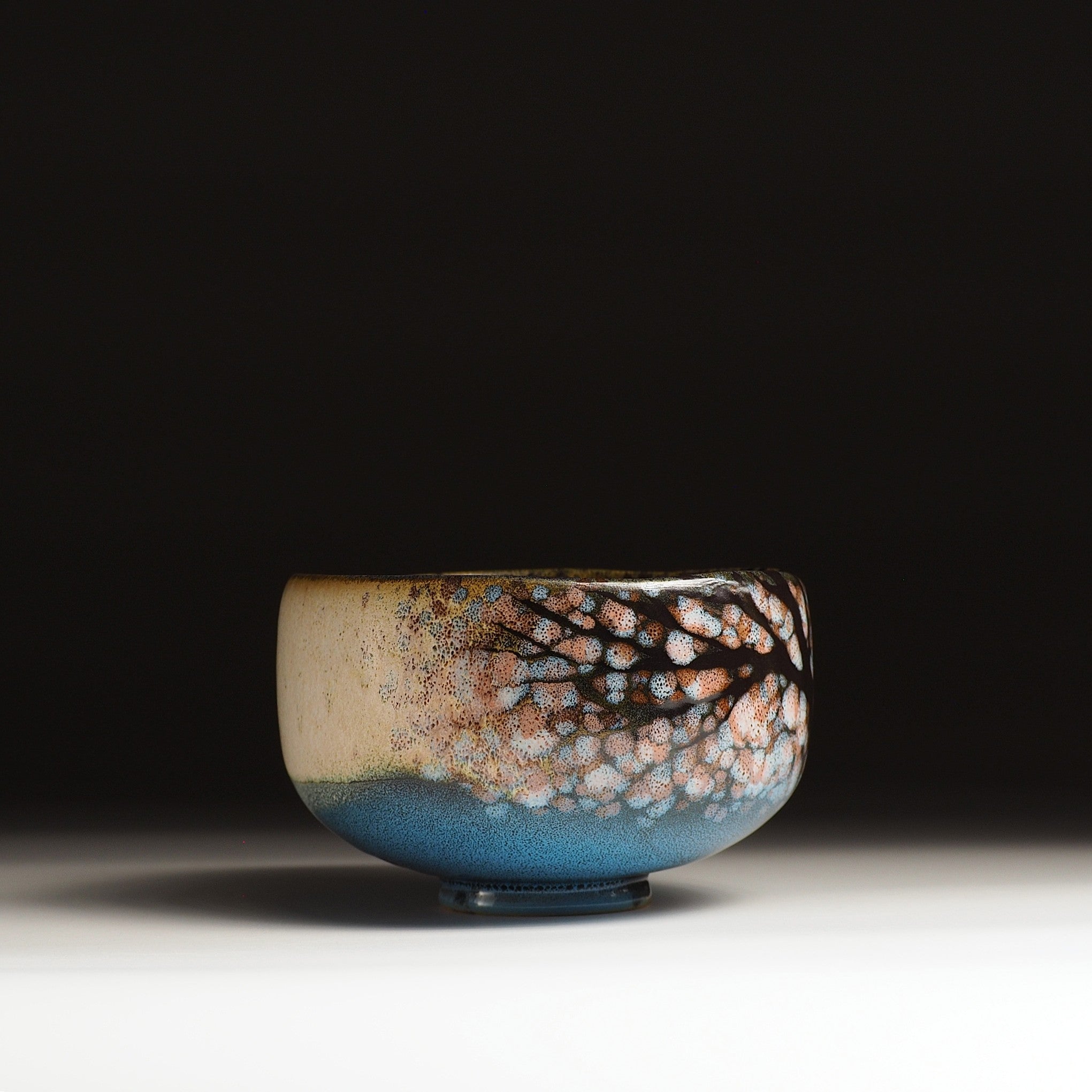 a side view of a Japanese ceramic matcha bowl designed with cherry blossoms and a tranquil lake against a black background. Tsukushi