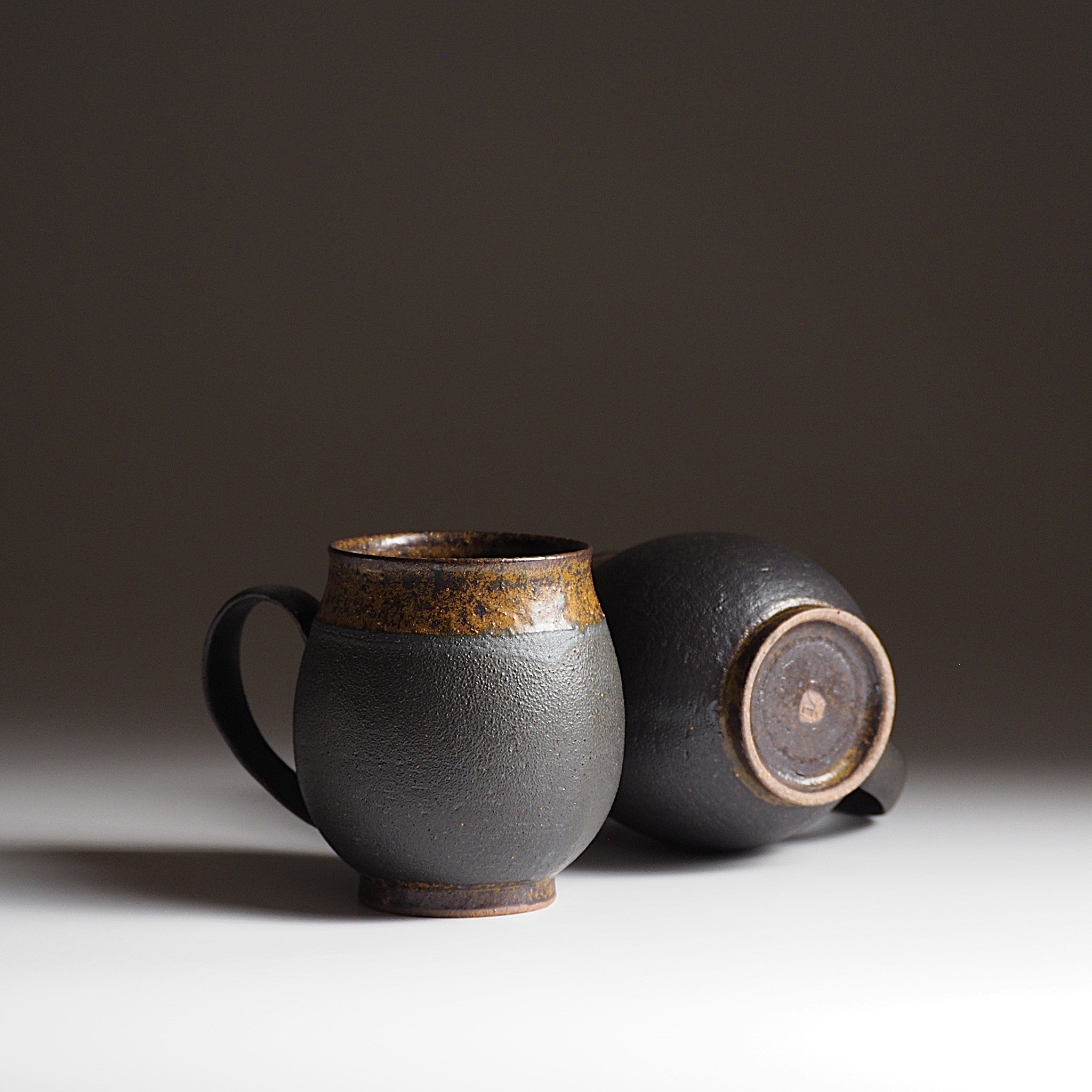 Two handcrafted black Japanese ceramic tea mugs on a white surface with a dark background, Tsukushi