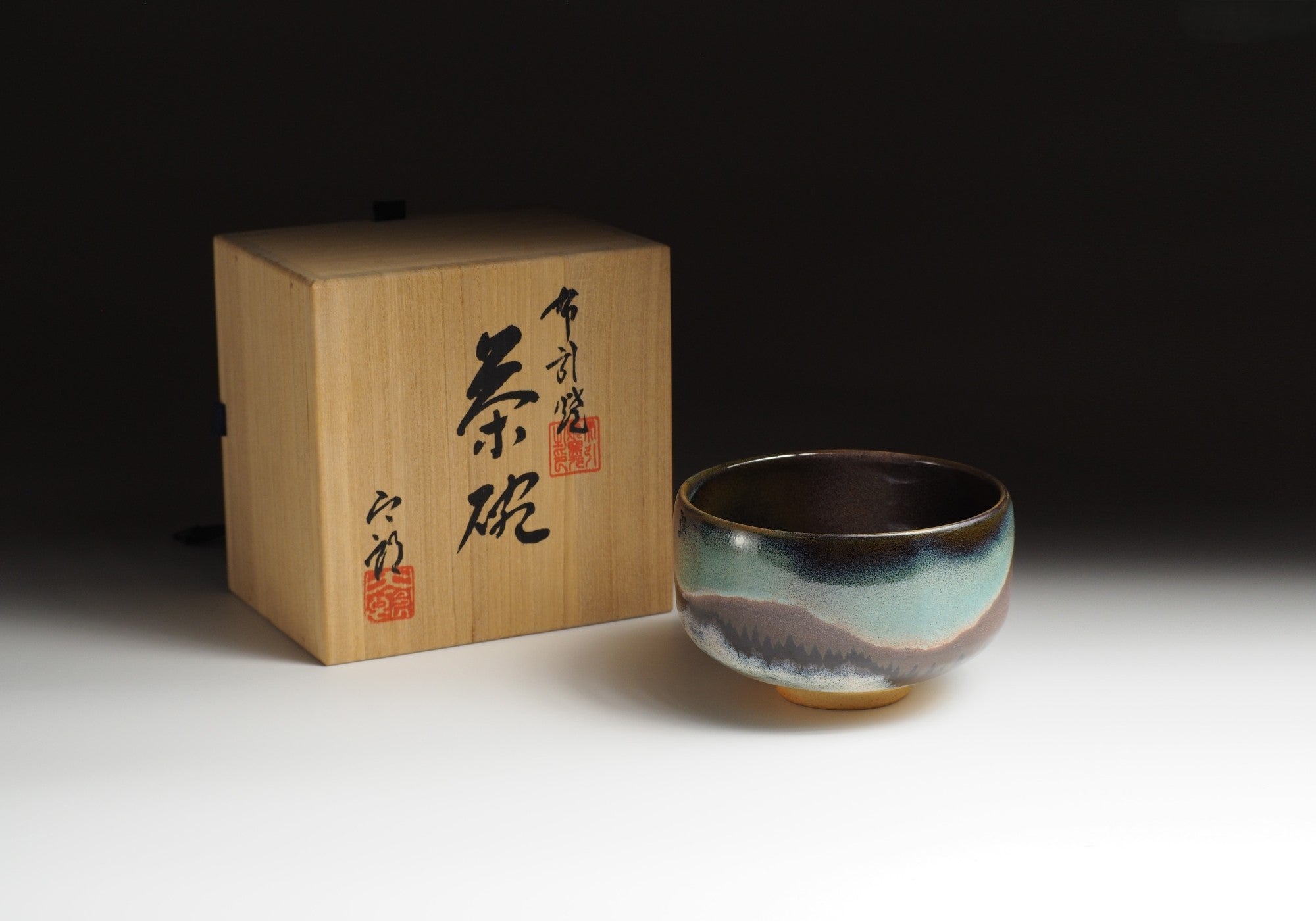 A Japanese ceramic matcha bowl with an Aurora sits next to a wooden box with calligraphy, set against a dark background. Tsukushi.