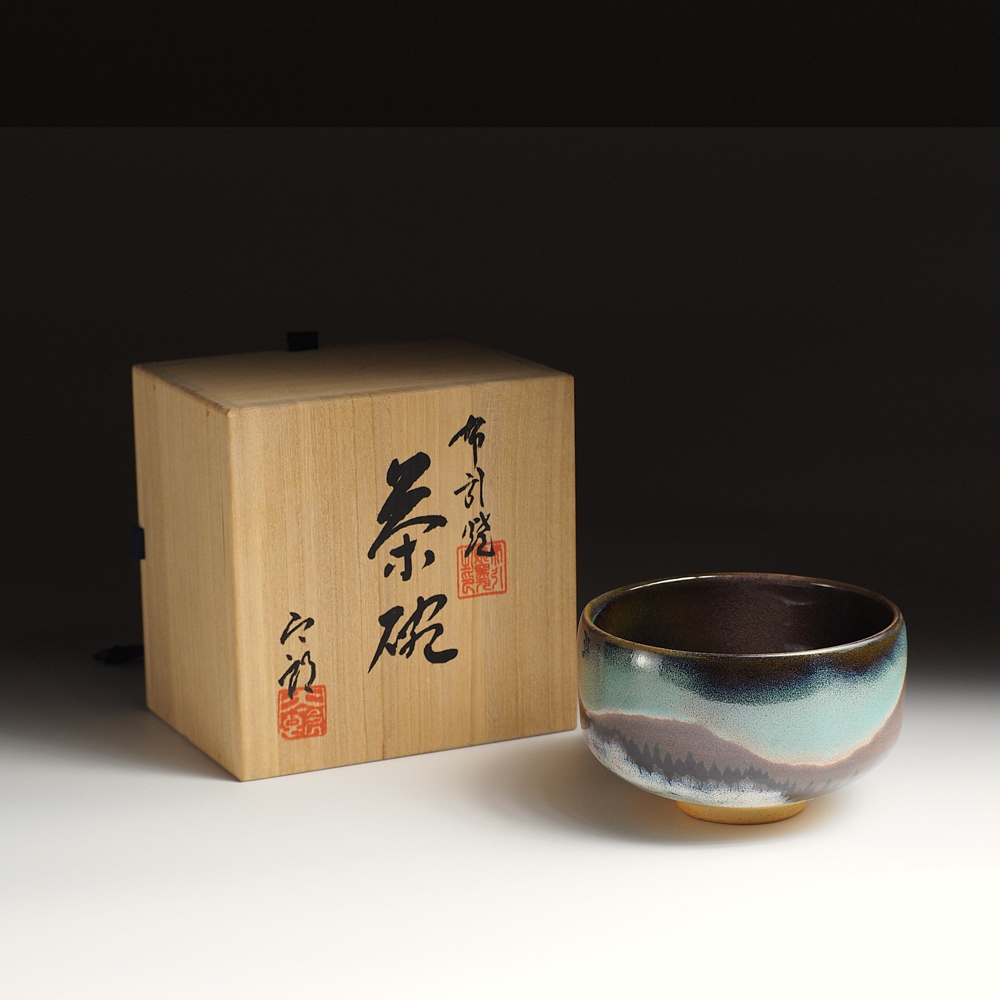 A Japanese ceramic matcha bowl with an aurora and a mountain sits next to a wooden box with calligraphy. Tsukushi