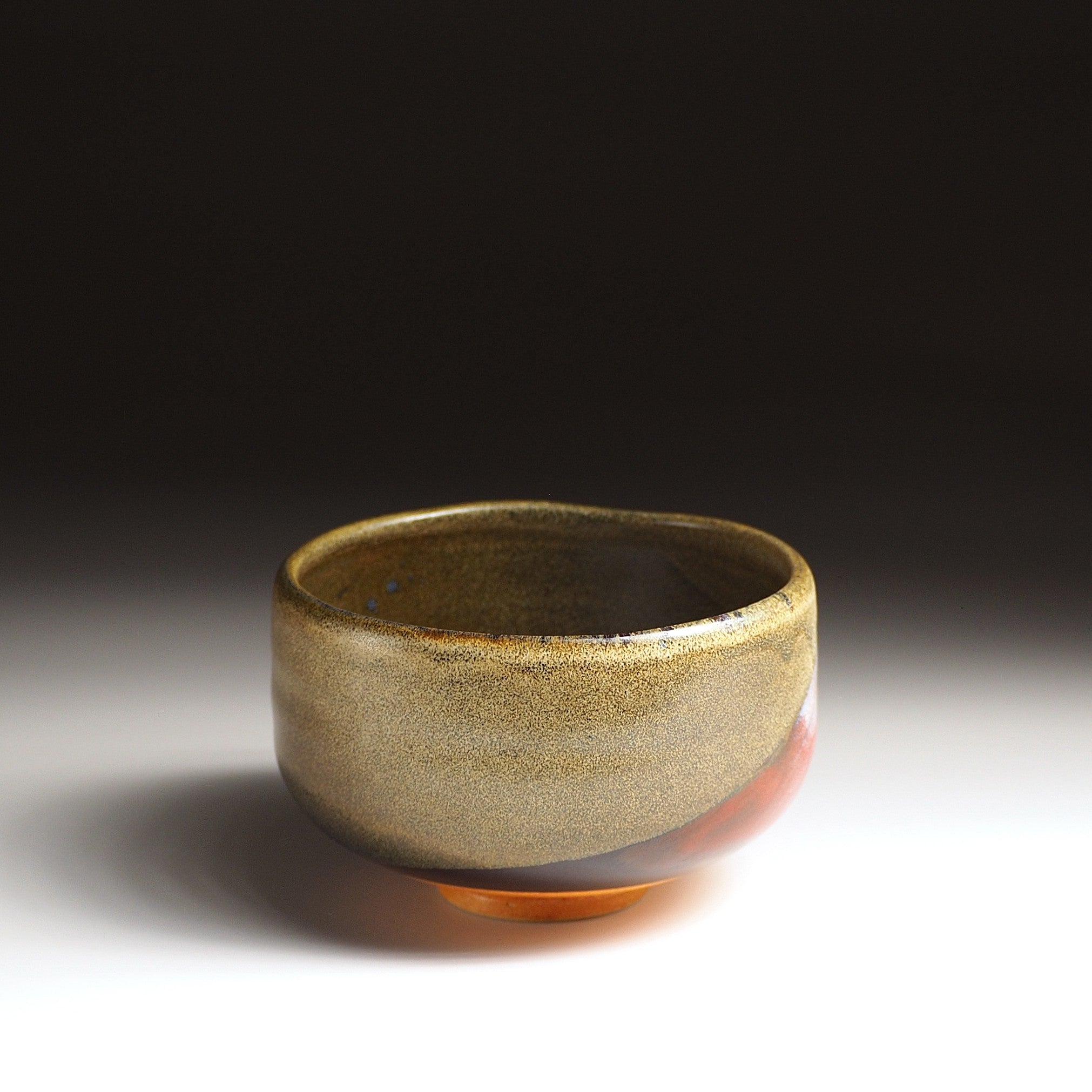 A side view of a Japanese ceramic matcha bowl adorned with Red Fuji on a brown surface, set against a black background. Tsukushi.