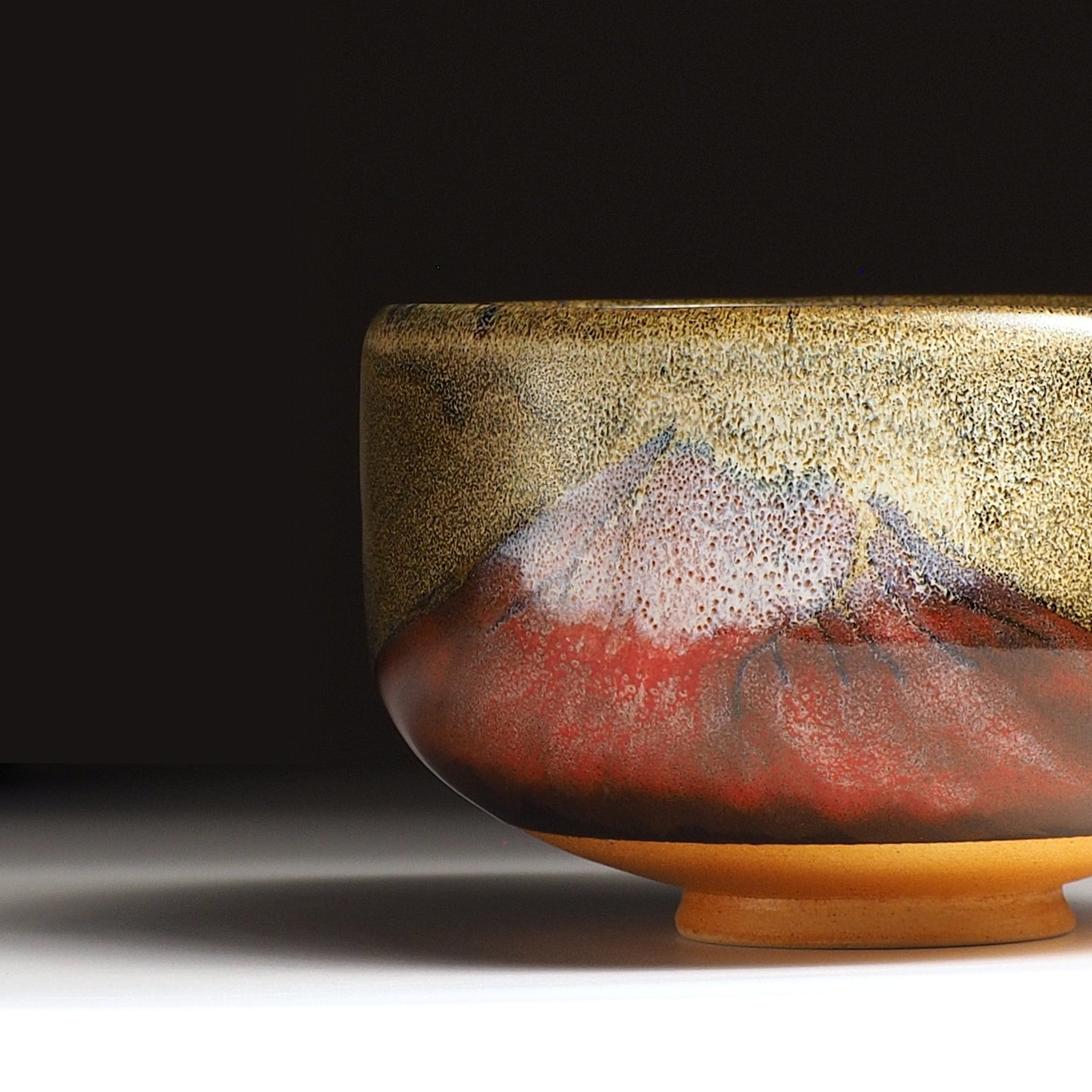 A close-up view of a Japanese ceramic matcha bowl adorned with Red Fuji on a brown surface, set against a black background. Tsukushi.