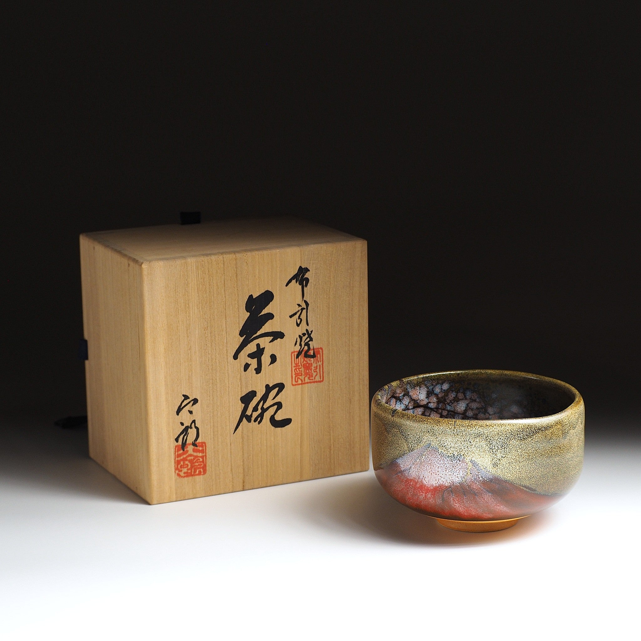 A Japanese ceramic matcha bowl depicting Red Fuji set against a dark background with wooden box.Tsukushi