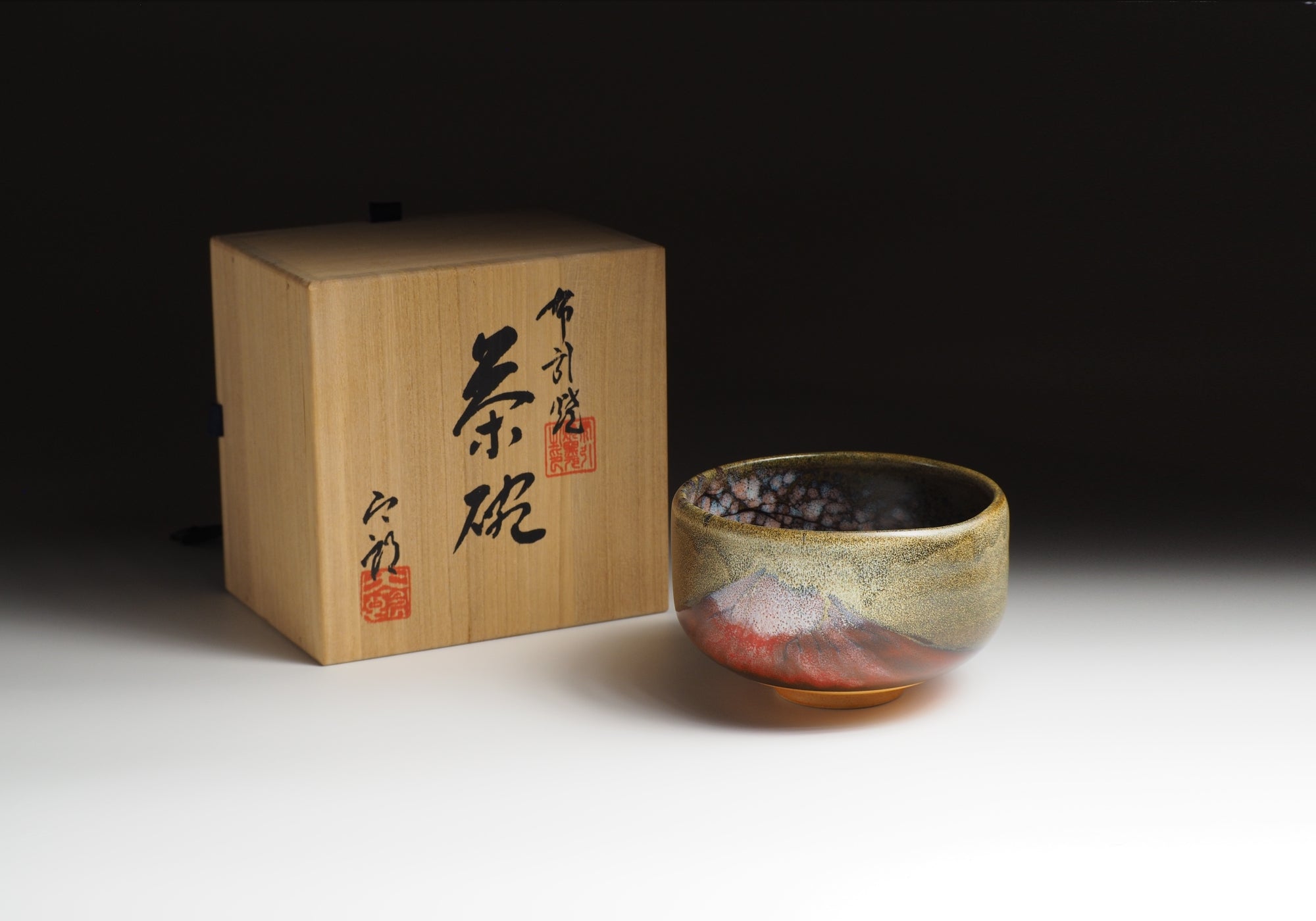 A Japanese ceramic matcha bowl with Mt. Red Fuji sits next to a wooden box with calligraphy. Tsukushi