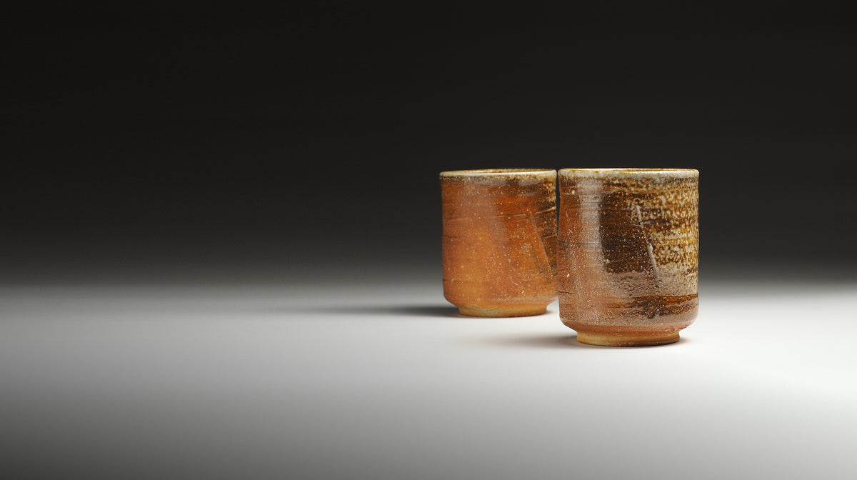 Two Shigaraki yunomi cups from with a textured, earthy brown and amber glaze, displayed against a dark gradient background.