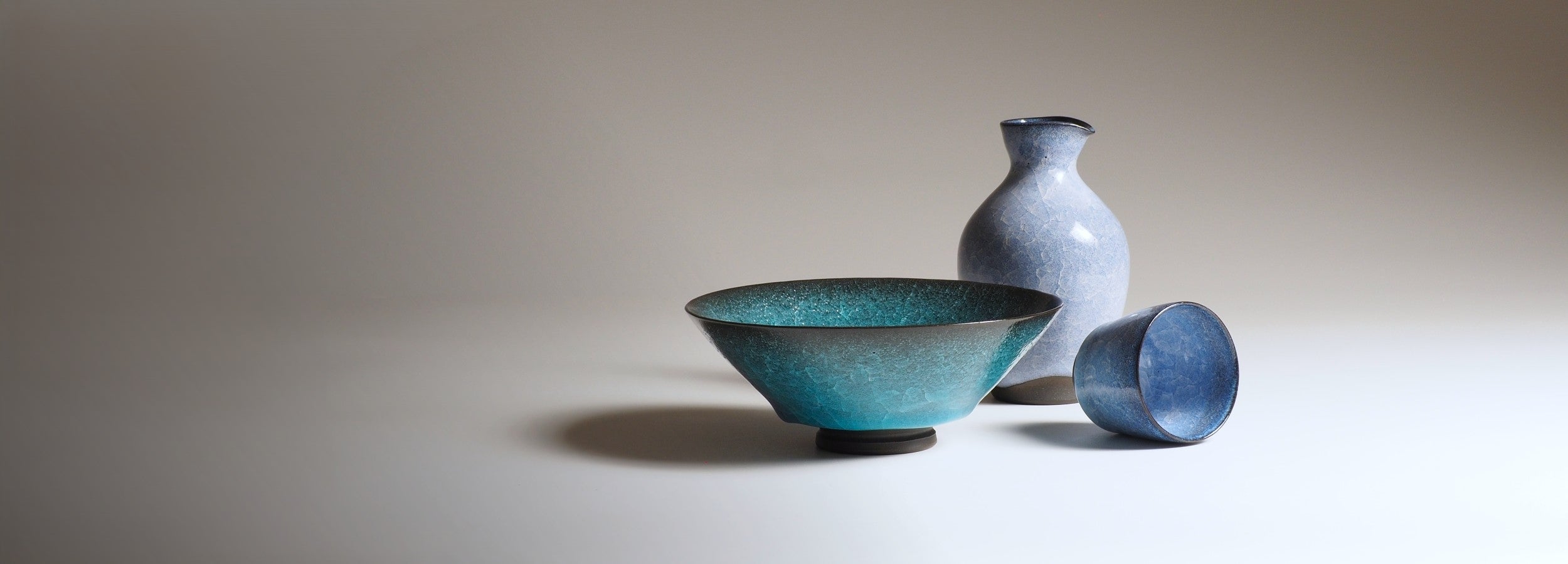 Three Japanese ceramics including a bowl, a tokkuri, and a sake cup on a beige background. Tsukushi