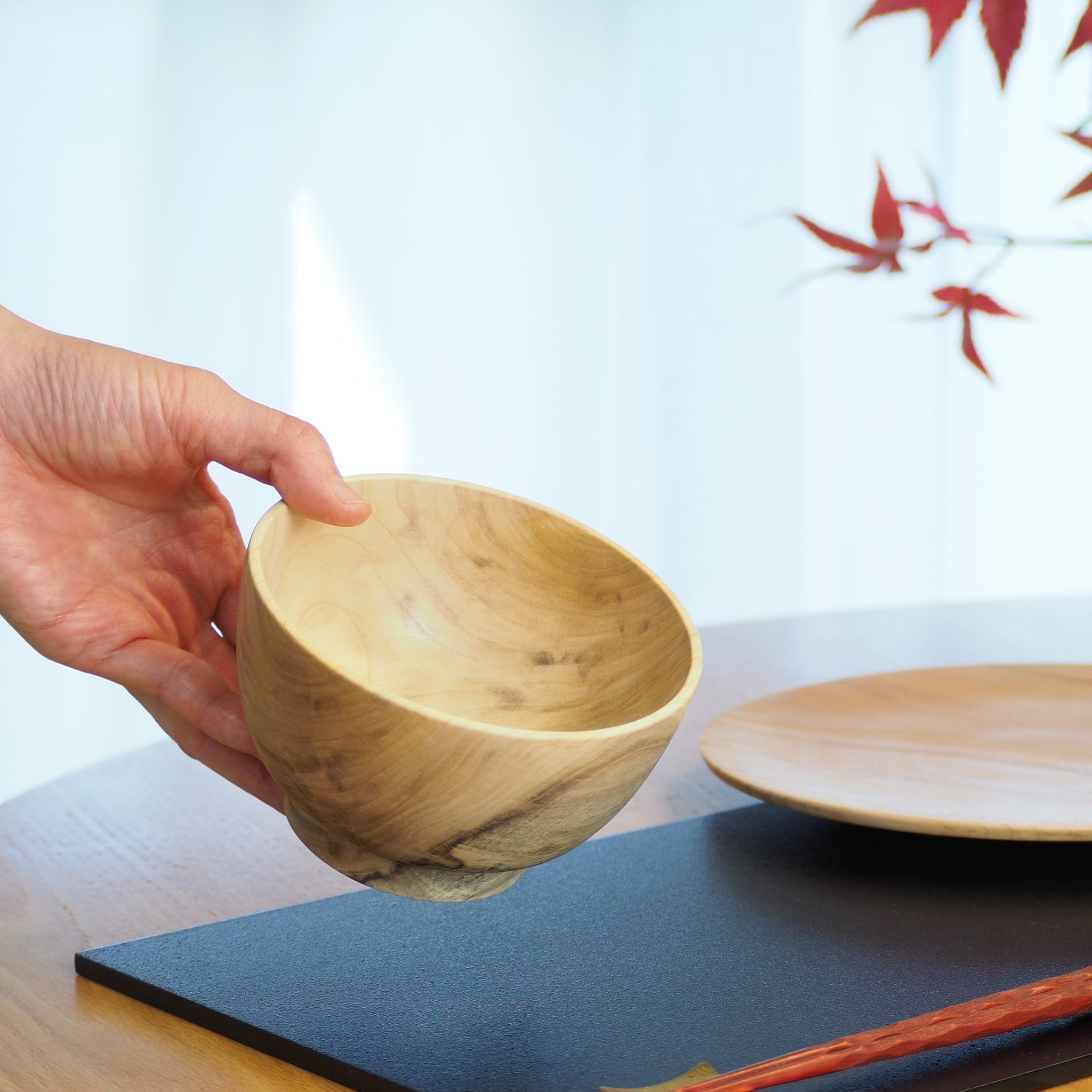 A hand holding a Japanese wooden bowl, with a blurred background featuring a red leaf design. Tsukushi