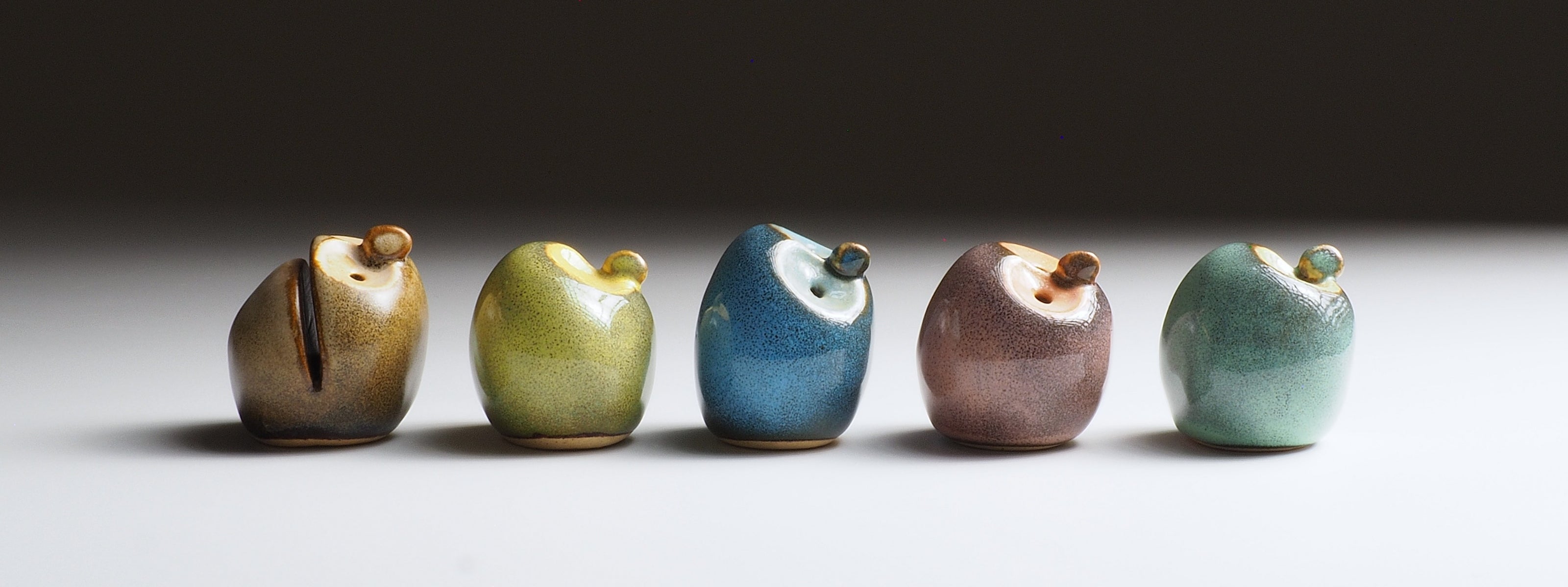 Five small ceramic owl-shaped figurines in assorted earthy glazes, arranged in a row on a white surface against a dark gray background.