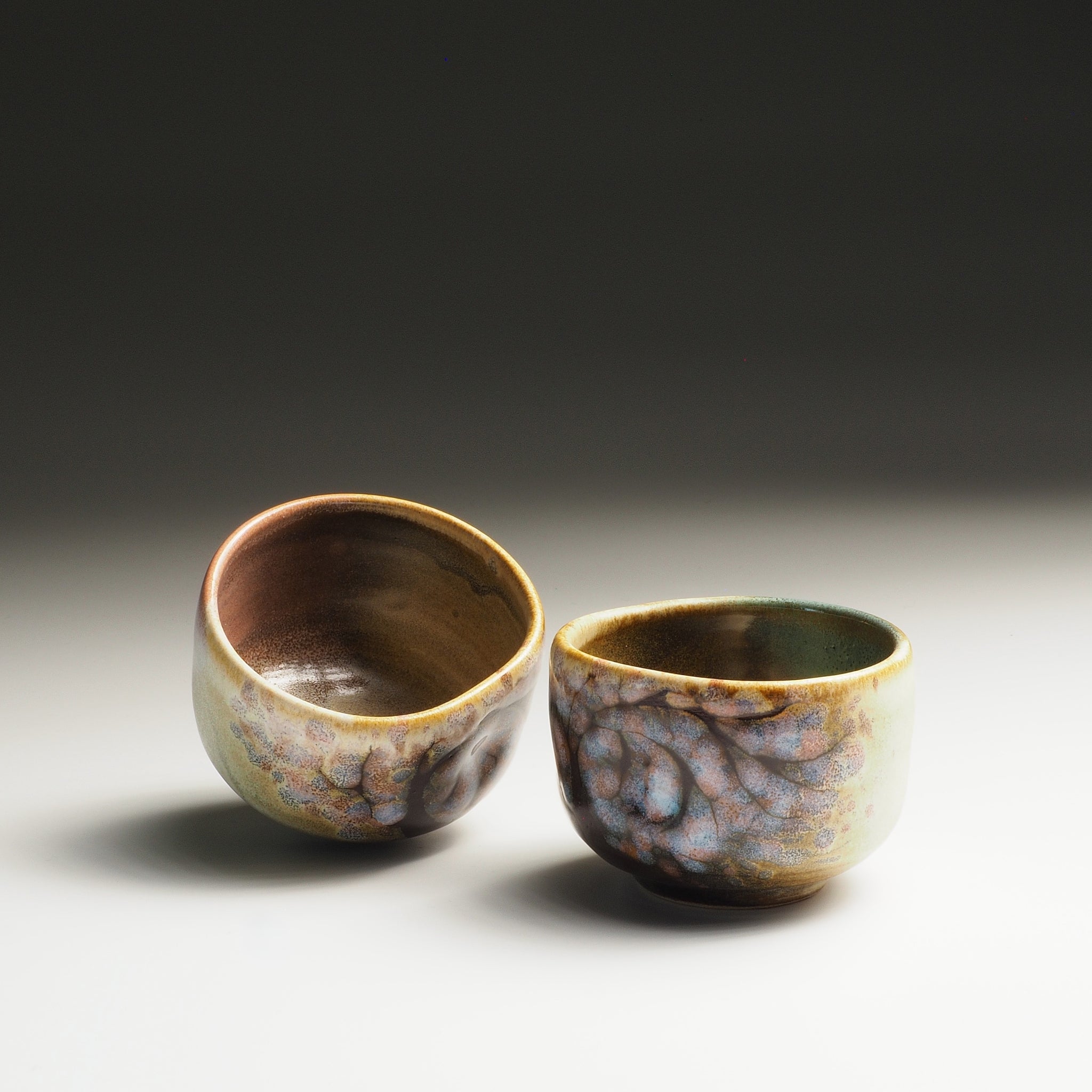 Two ceramic guinomi sake cups with sakura decorations on a gradient background