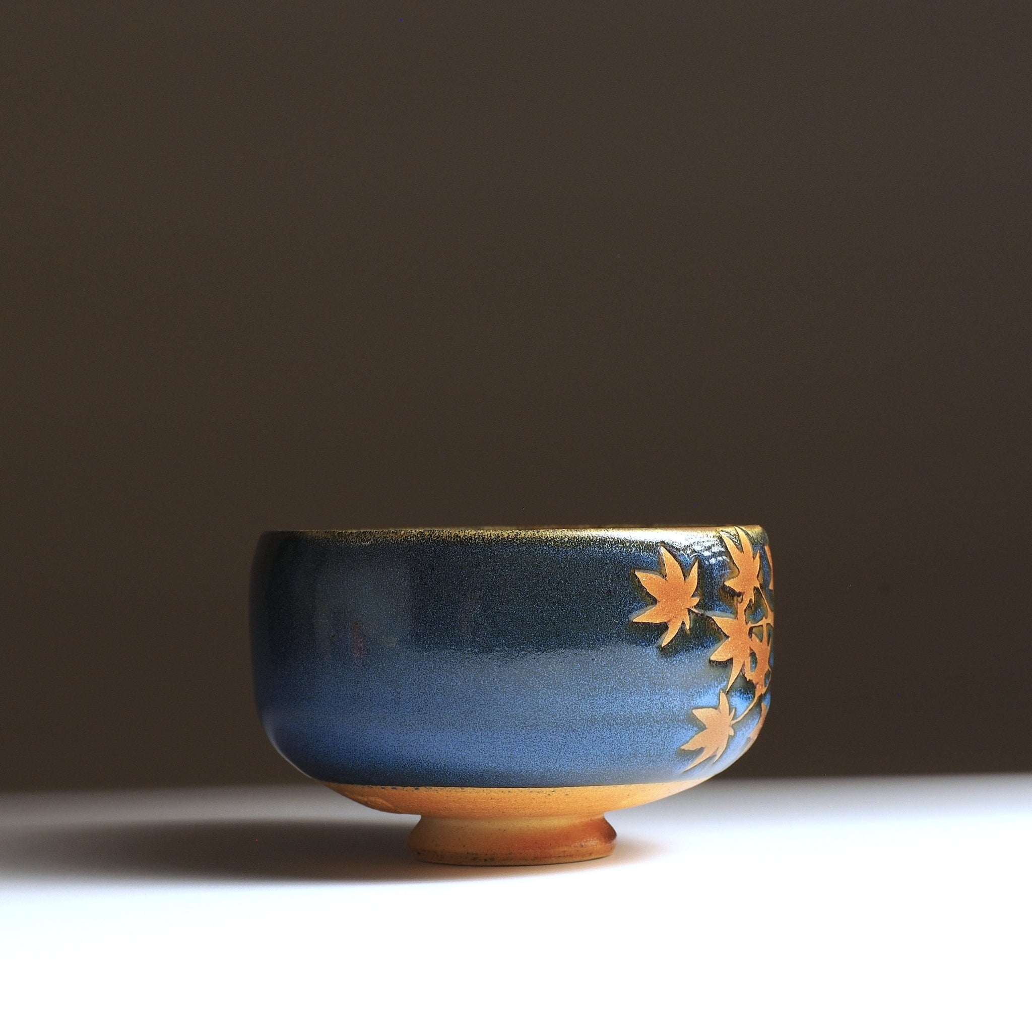 Sōshū matcha bowl, side view, sparse momiji leaves in natural clay against a gradient blue Nanasai Tenmoku glaze, unglazed amber foot ring visible