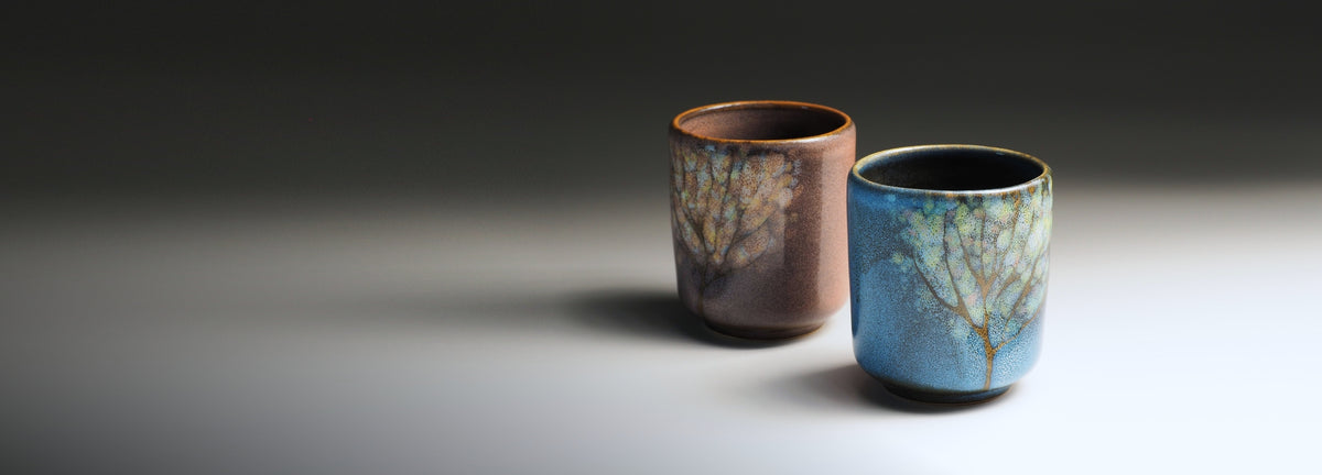 Two Japanese ceramic cups with tree-like glaze patterns, one in soft pink and the other in blue, set against a dark-to-light gray gradient background.