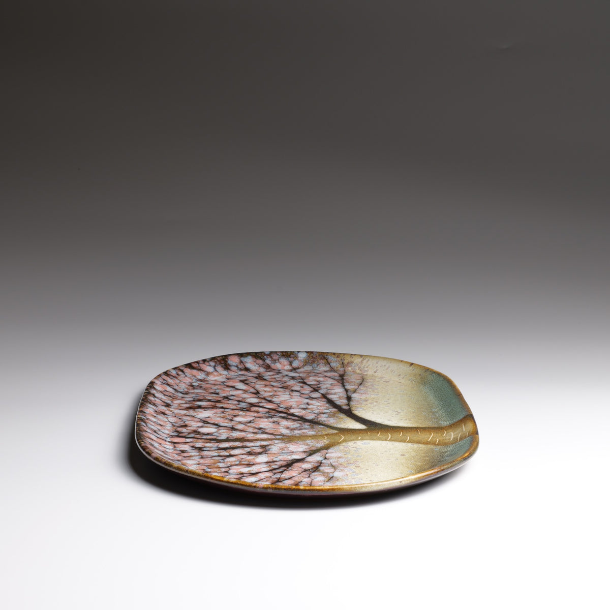 Hanami handmade Japanese ceramic plate with pink cherry blossoms, a golden tree trunk, and soft green glaze, shown from a low front angle.