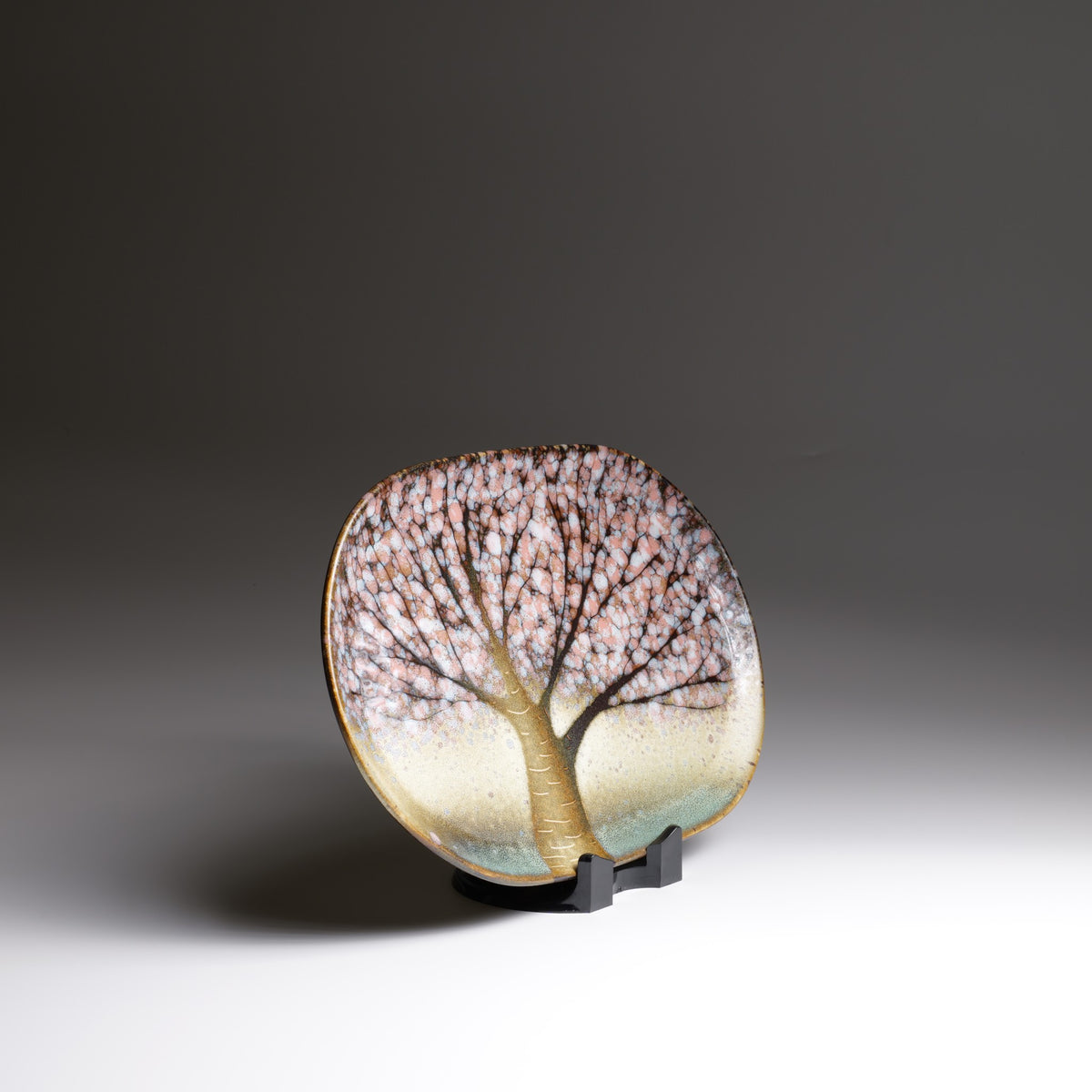 Front view of the Hanami handmade Japanese ceramic plate on a black stand, featuring pink cherry blossoms, golden branches, and soft green glaze.