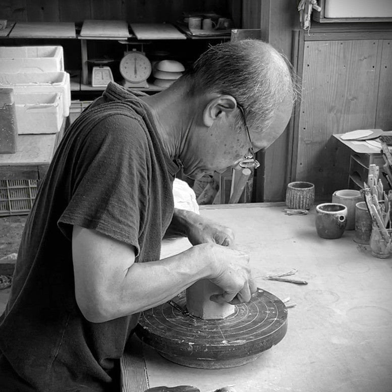 A black and white photograph of Hitoshi Minagawa shaping a ceramic pot on a pottery wheel in a workshop.
