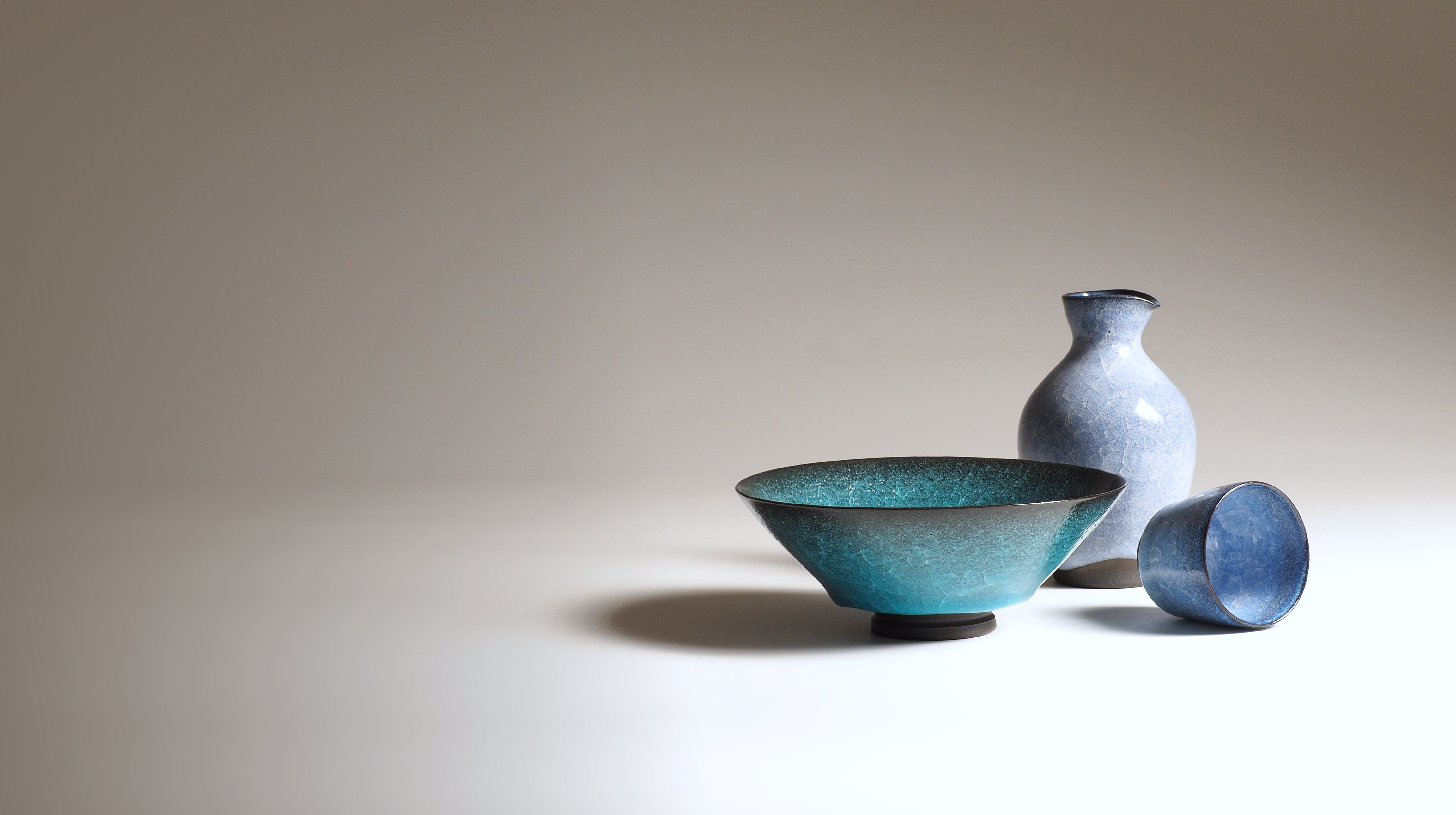 Three Japanese ceramic pieces including a bowl, a vase, and a sake cup on a light background. Tsukushi
