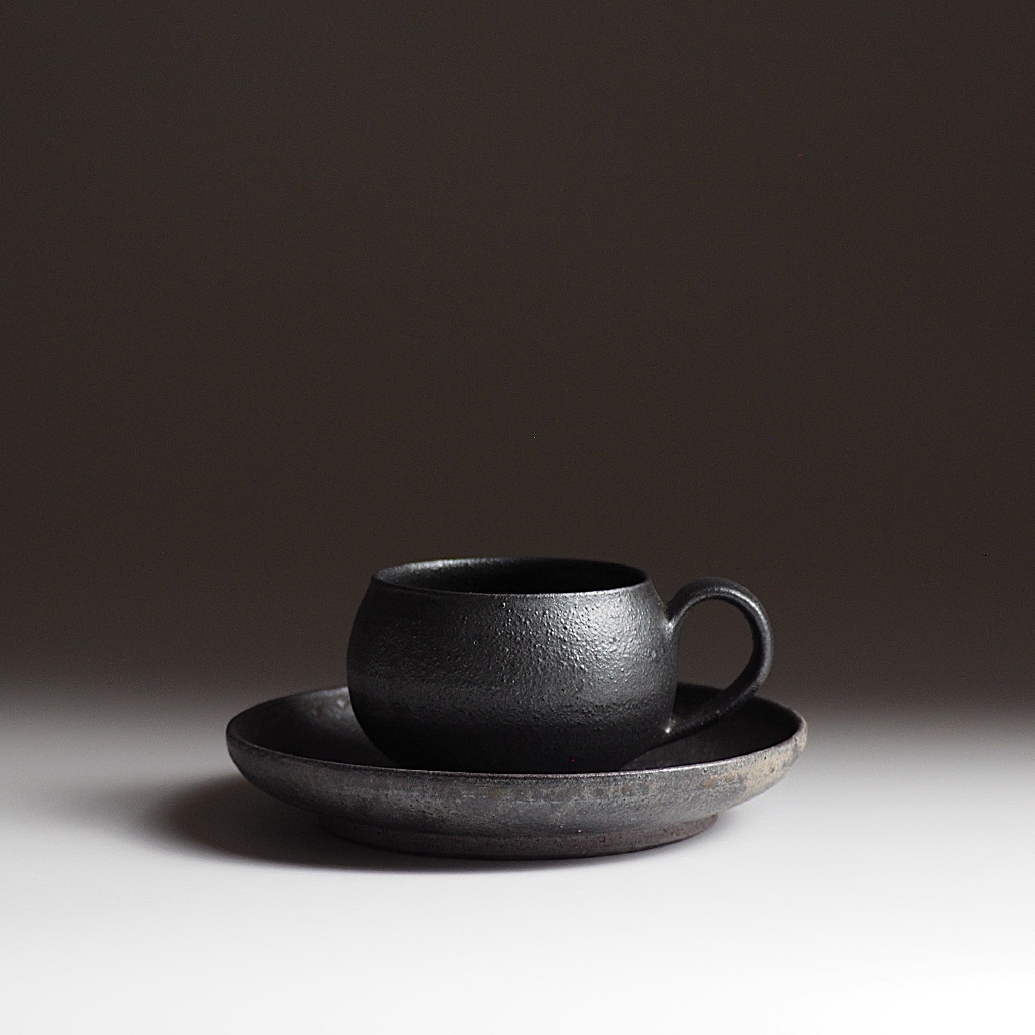 Side view of handcrafted black Japanese ceramic espresso cup and saucer set on a light gray background, Tsukushi