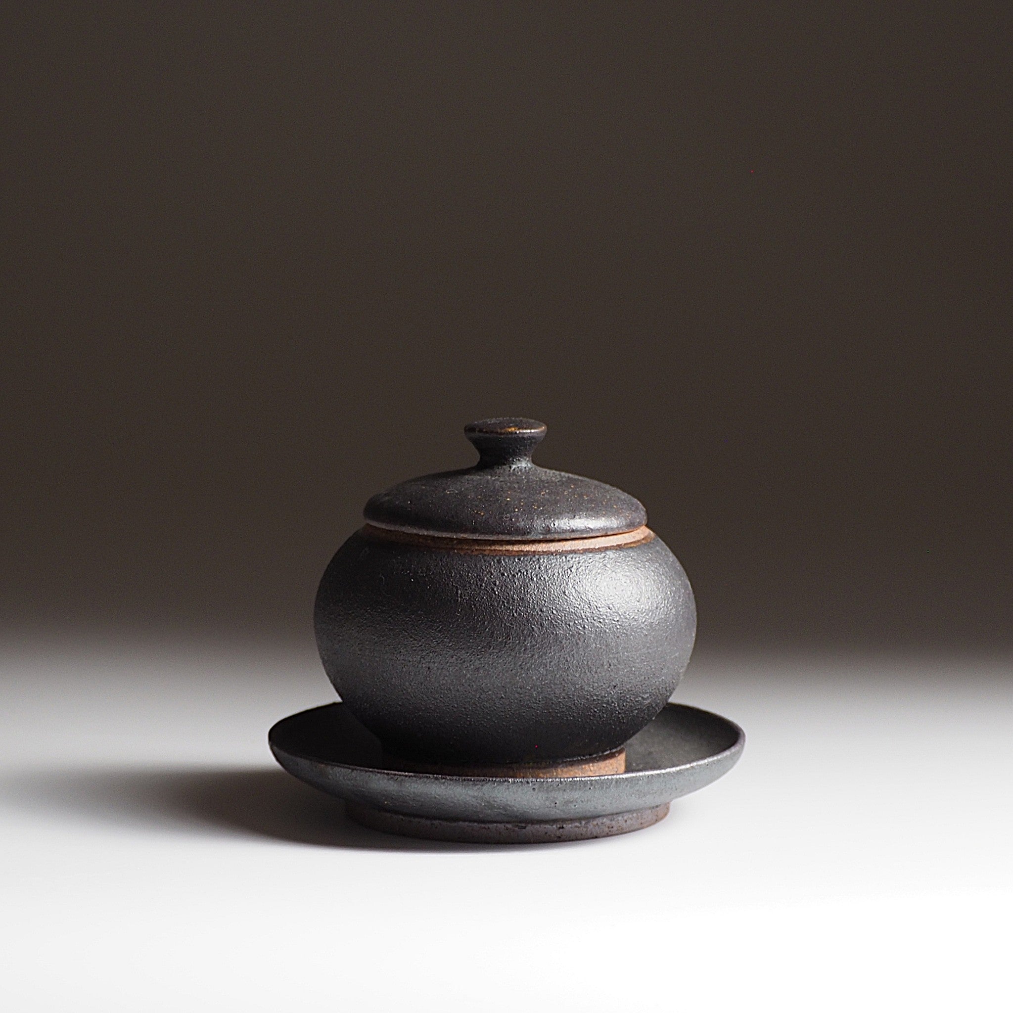 Black Japanese ceramic chawanmusi bowl with lid and matching saucer on a dark background. Tsukushi