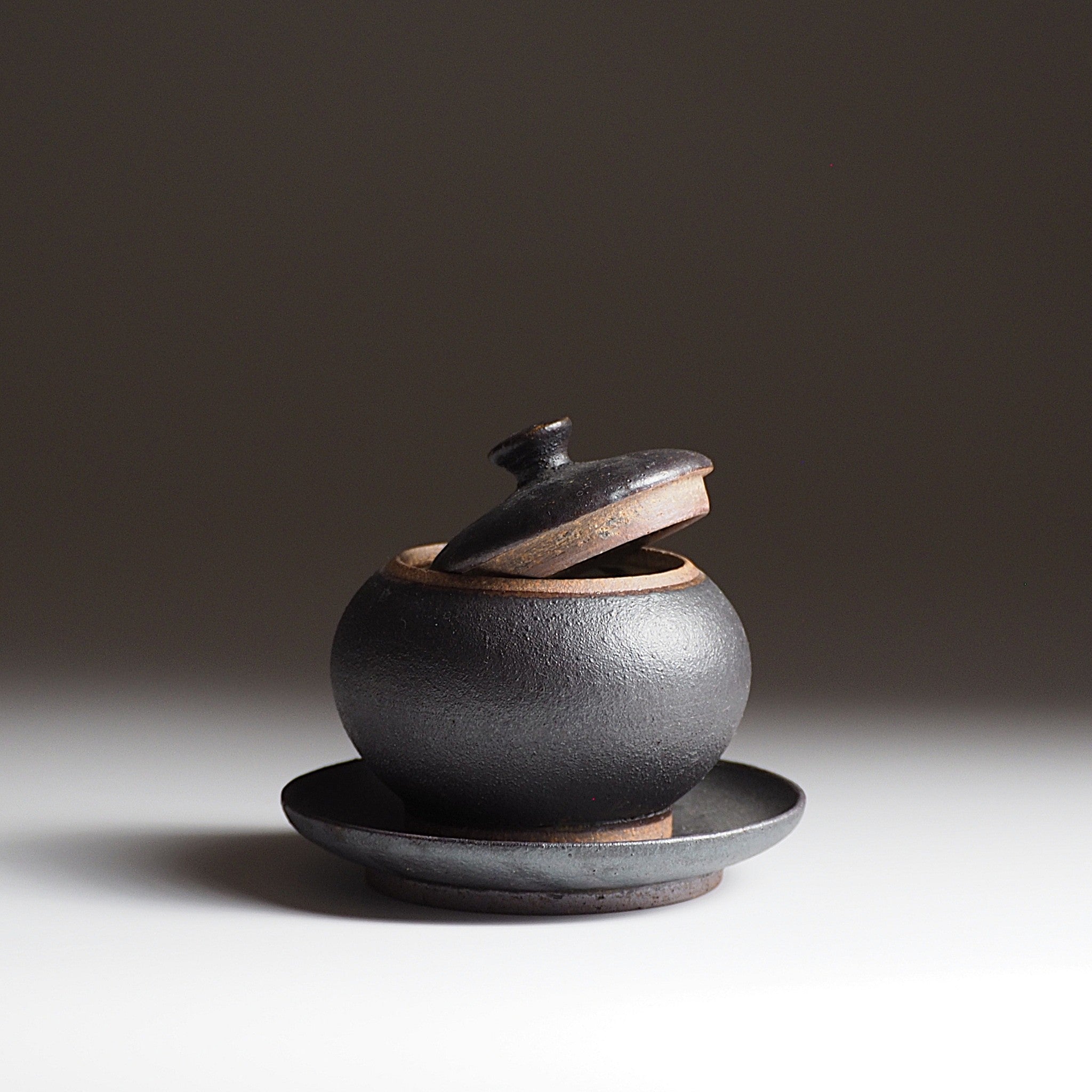 Black Japanese pottery chawanmusi bowl with matching saucer against a dark background. Tsukushi