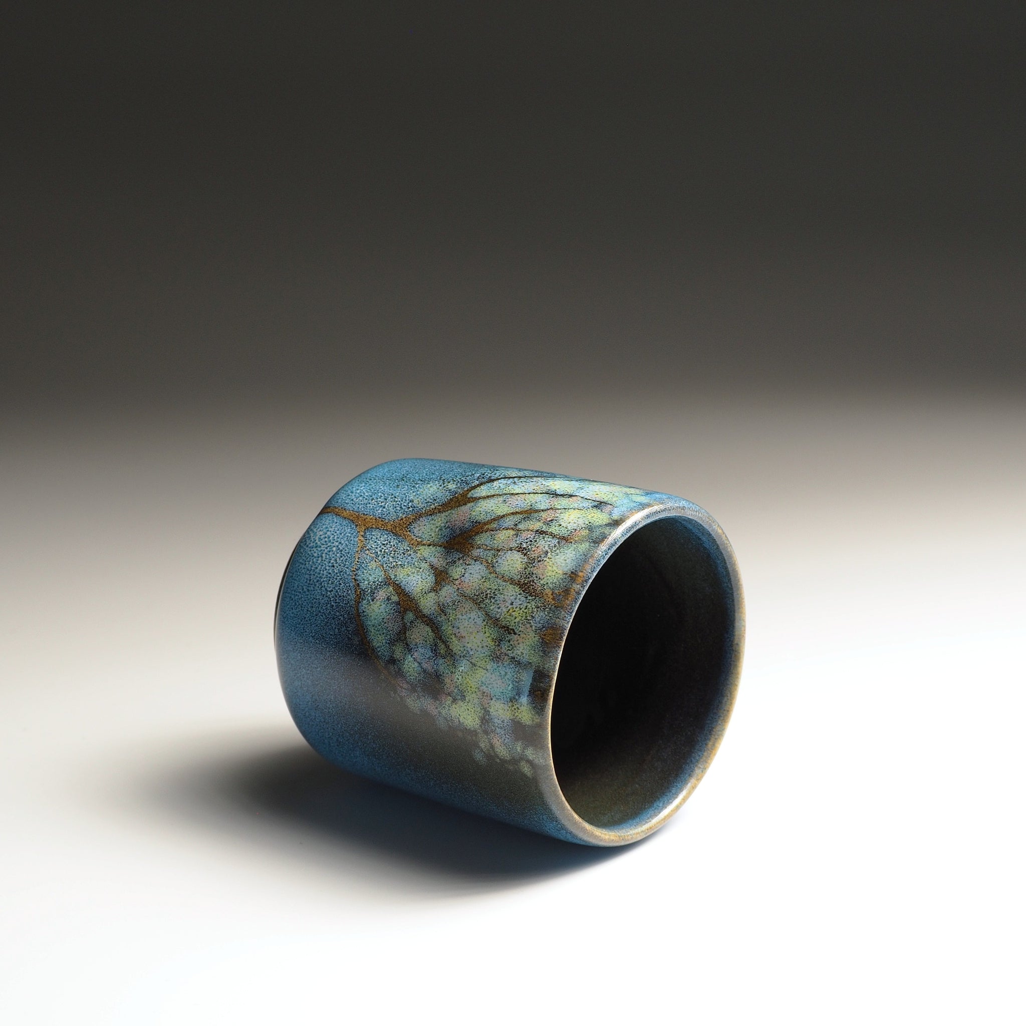 Ceramic yunomi teacup with blue glaze and leaf pattern on a gradient background. Tsukushi