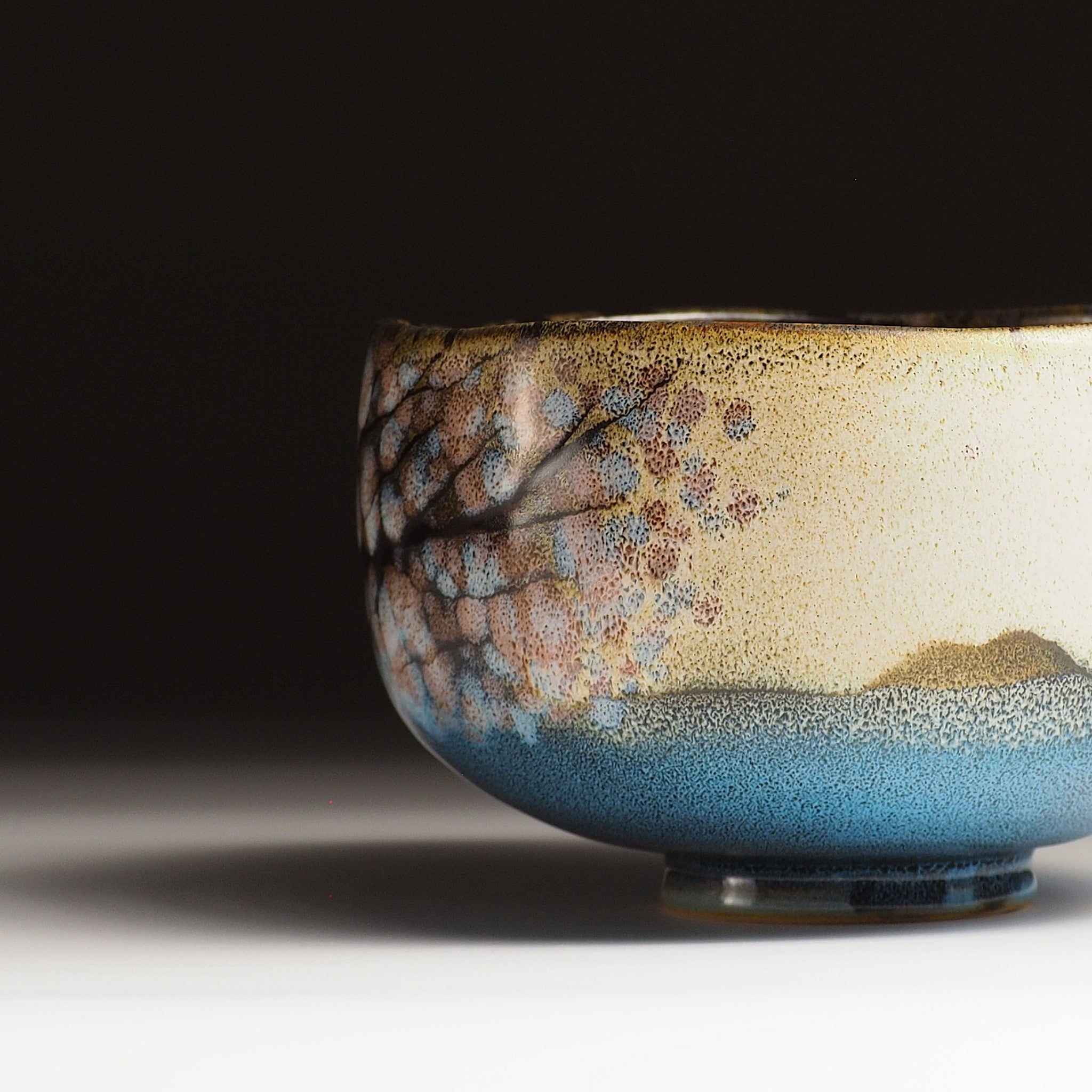 A close-up of a Japanese ceramic matcha bowl designed with cherry blossoms and a still lake against a black background. Tsukushi