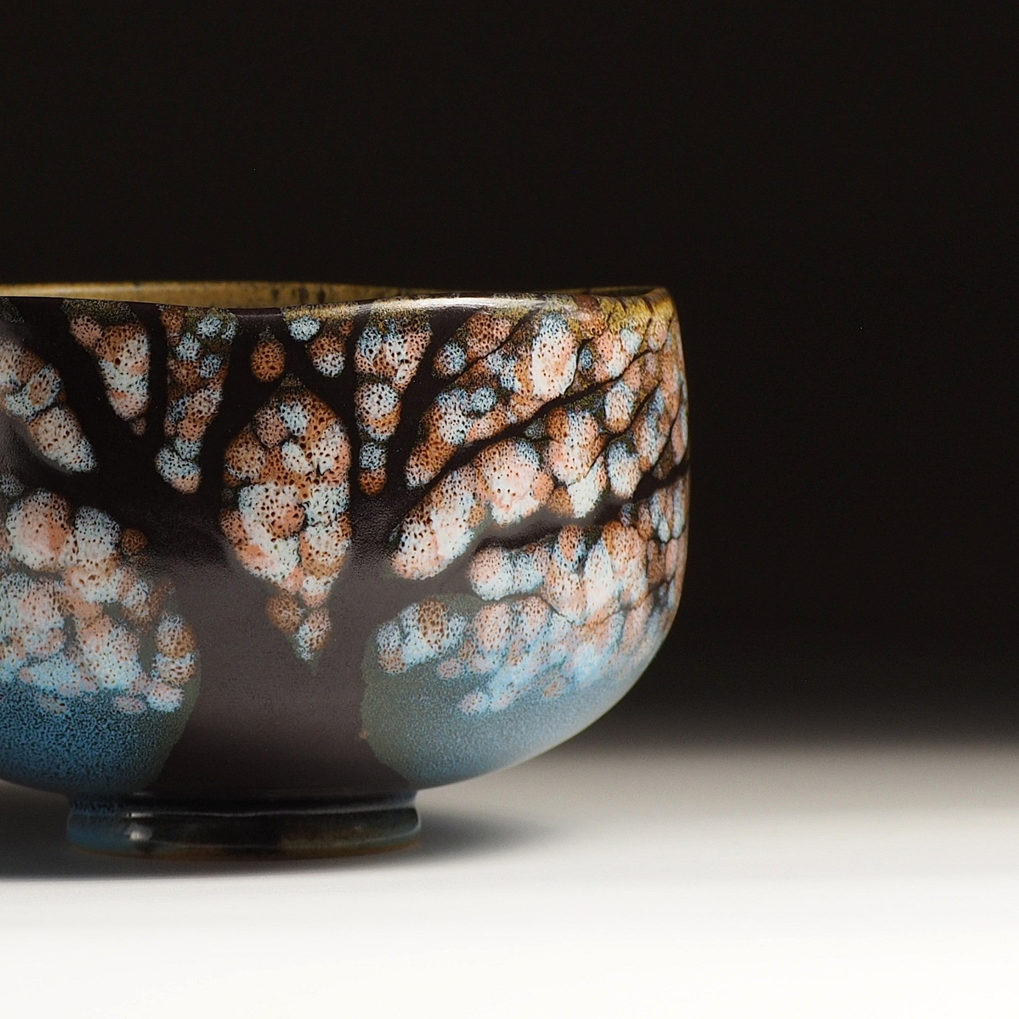 A close-up of a Japanese ceramic matcha bowl designed with cherry blossoms and a serene lake against a black background. Tsukushi