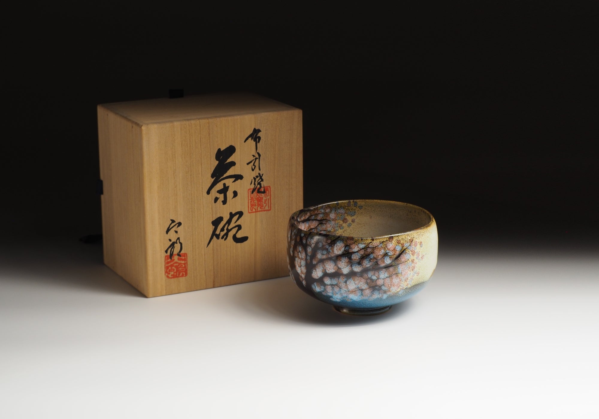 A Japanese ceramic matcha bowl with cherry blossoms sit next to a wooden box with calligraphy. Tsukushi