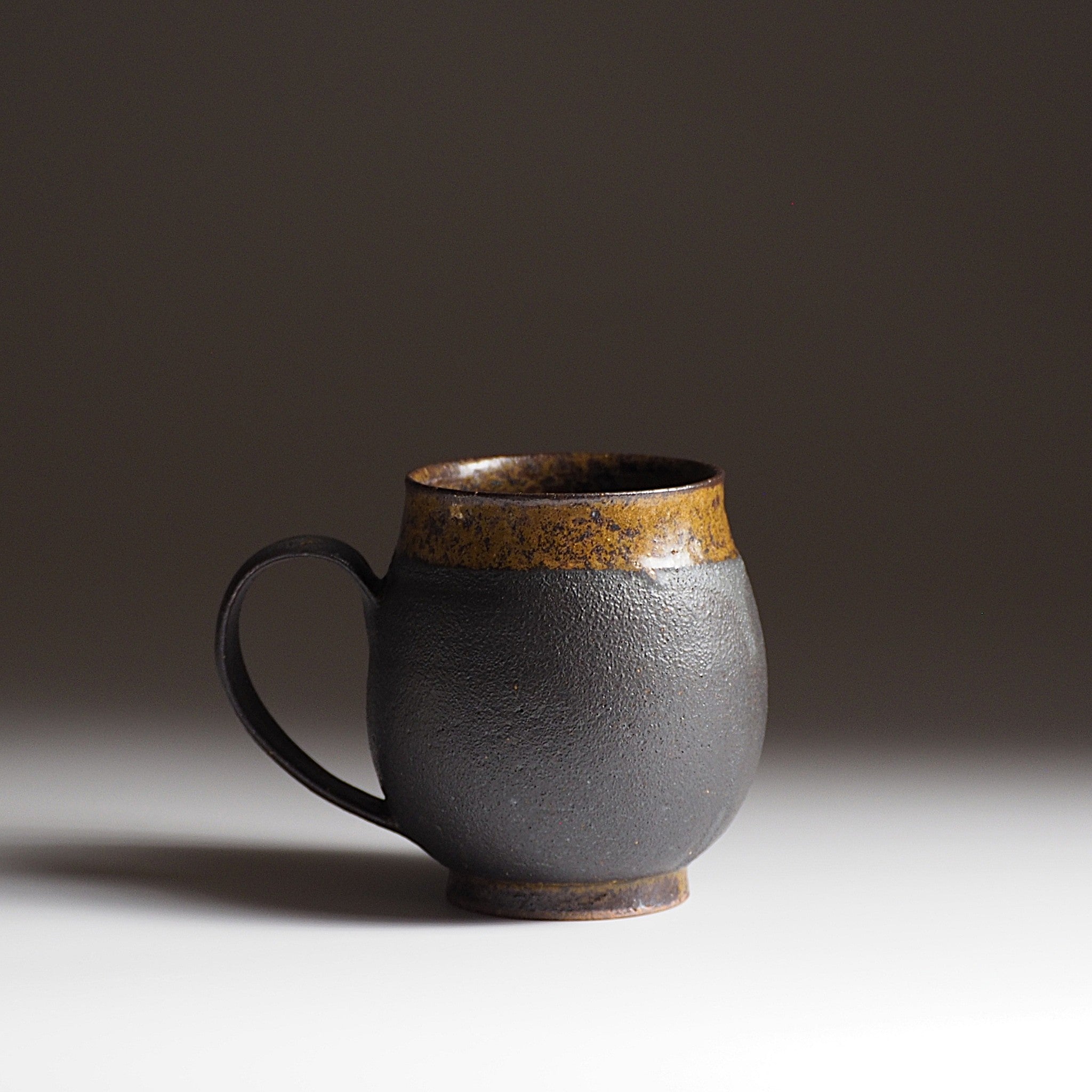 Side view of  handcrafted black Japanese ceramic tea mug on a white surface with a dark background, Tsukush