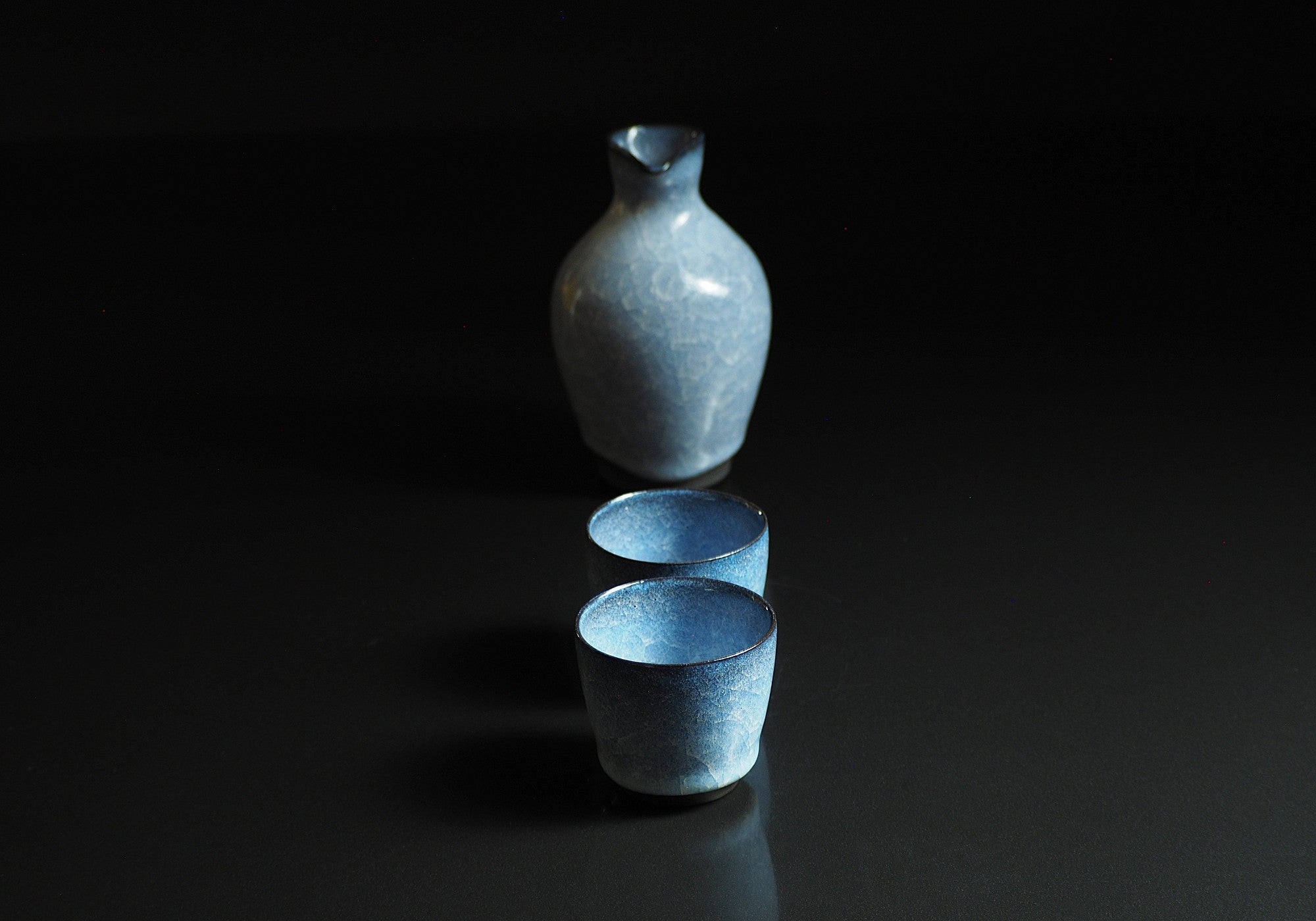 Handcrafted crazing Japanese ceramic Tokkuri and sake cups black background with reflection, Tsukushi
