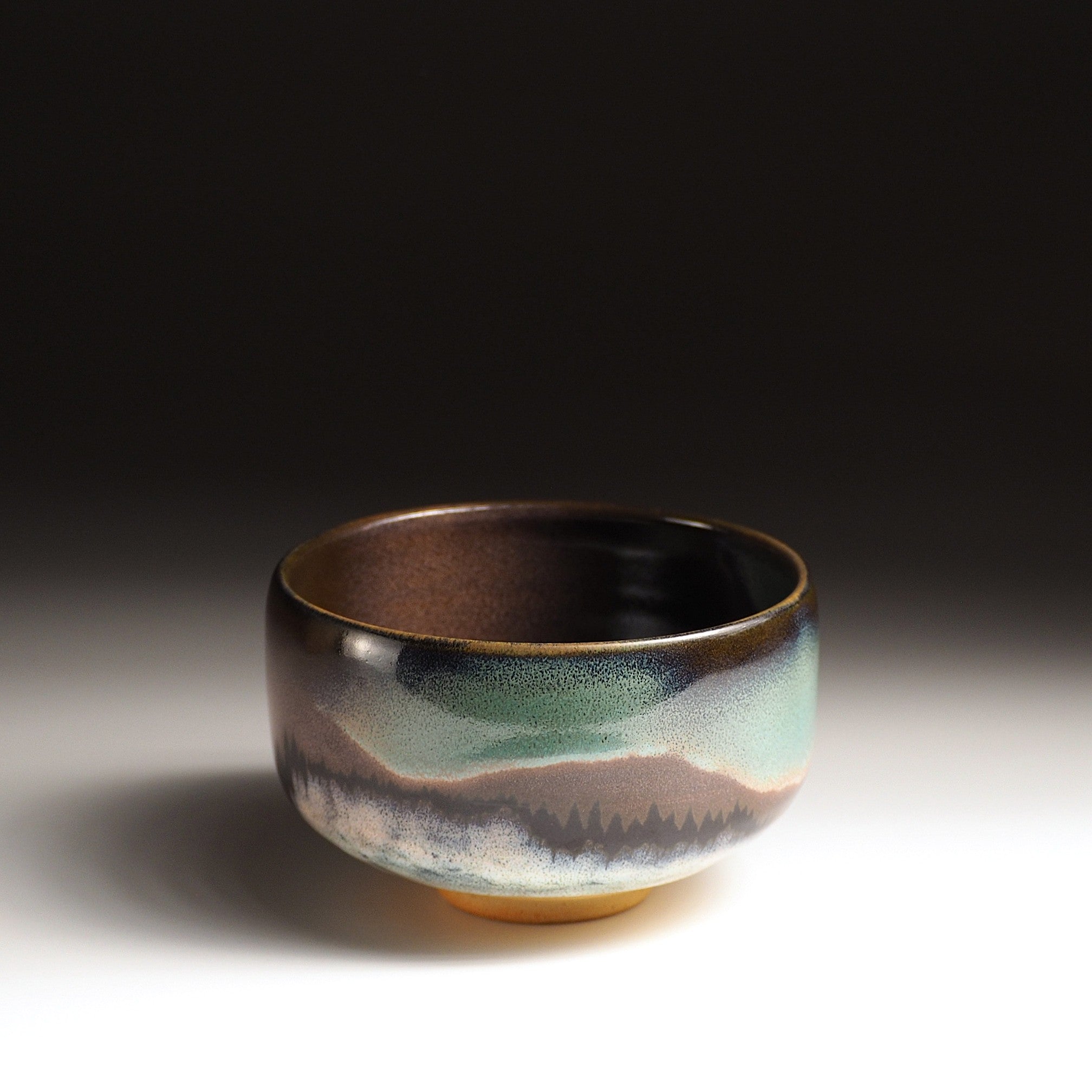 A Japanese ceramic matcha bowl designed with an aurora, a mountain, and a grove of trees, set against a black background. Tsukushi