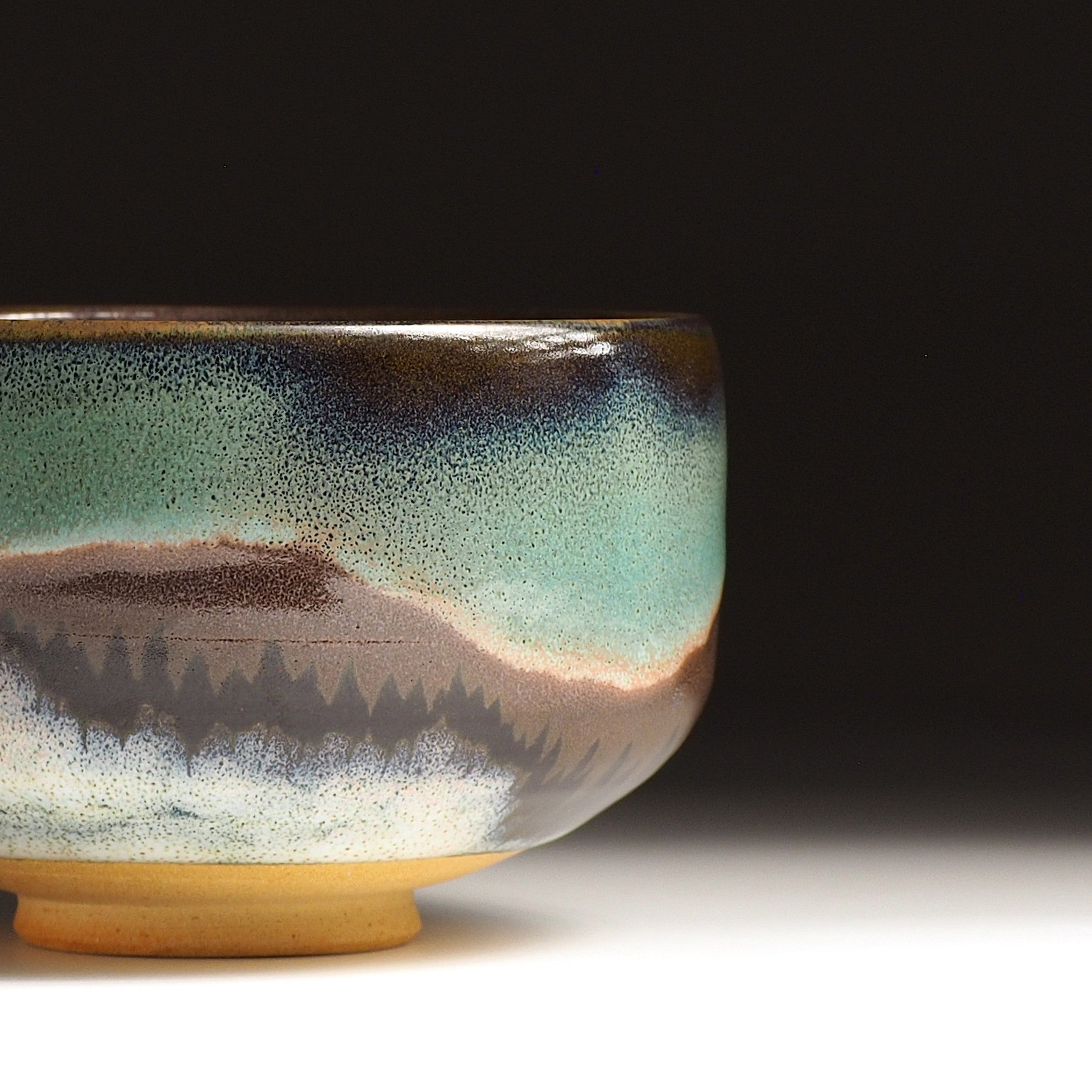 A Japanese ceramic matcha bowl adorned with an aurora, a mountain, and a grove of trees, set against a black background. Tsukushi