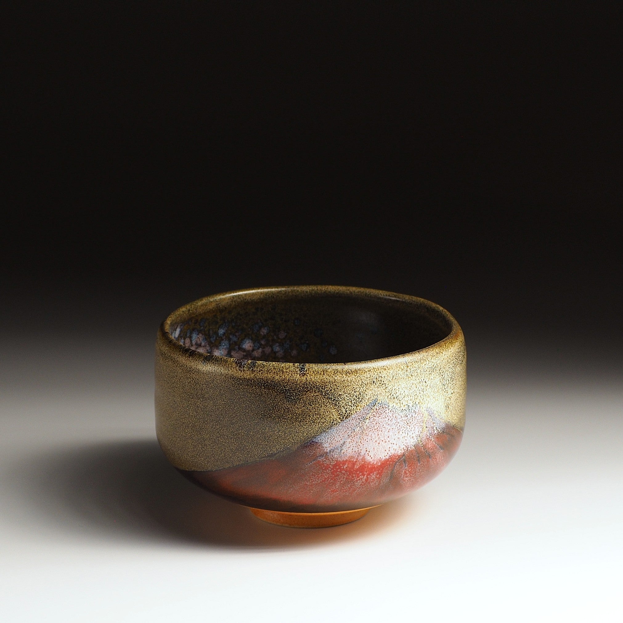 A Japanese ceramic matcha bowl adorned with Red Fuji a brown surface, set against a black background. Tsukushi.