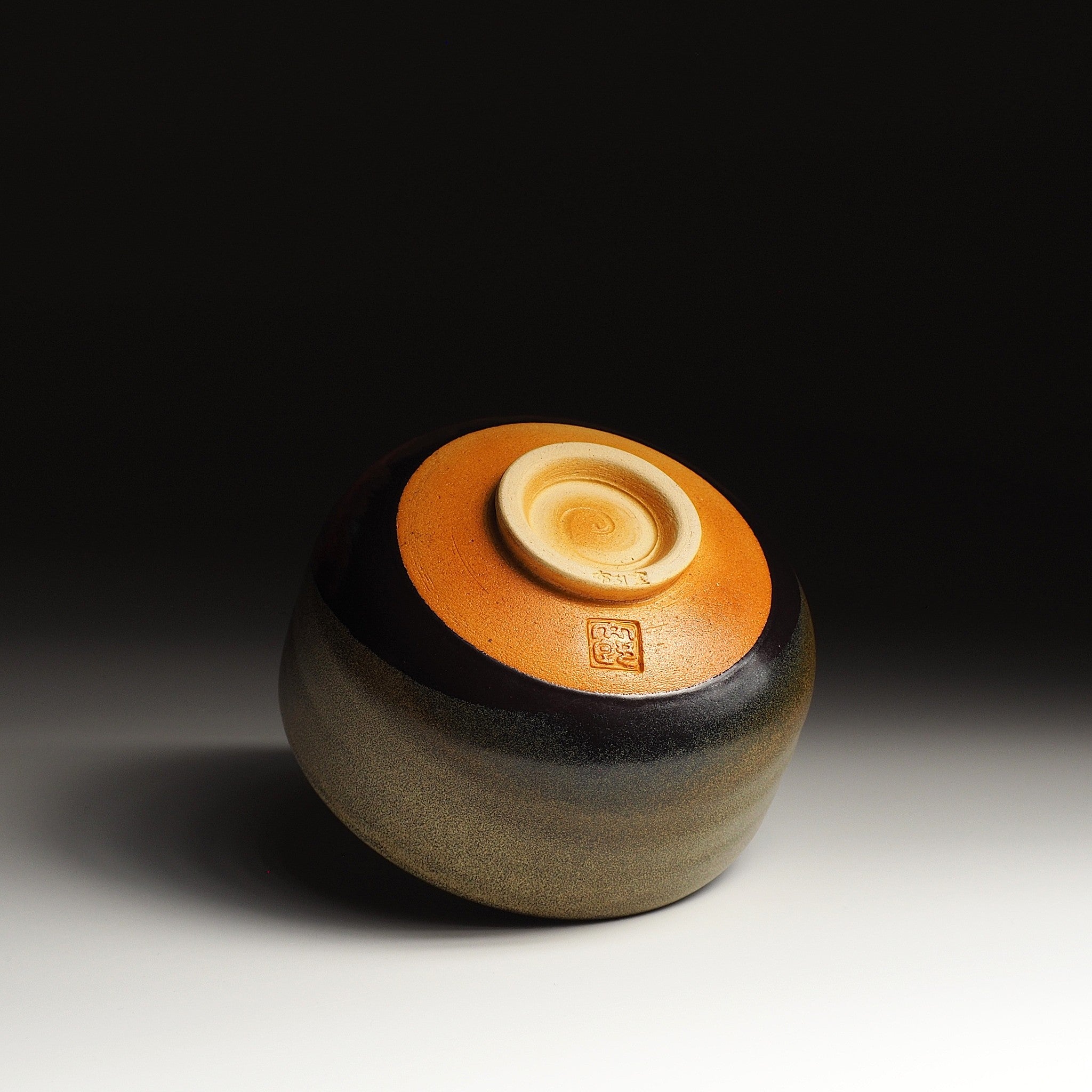 A bottom view of a Japanese ceramic matcha bowl adorned with Red Fuji on a brown surface, set against a black background. Tsukushi.