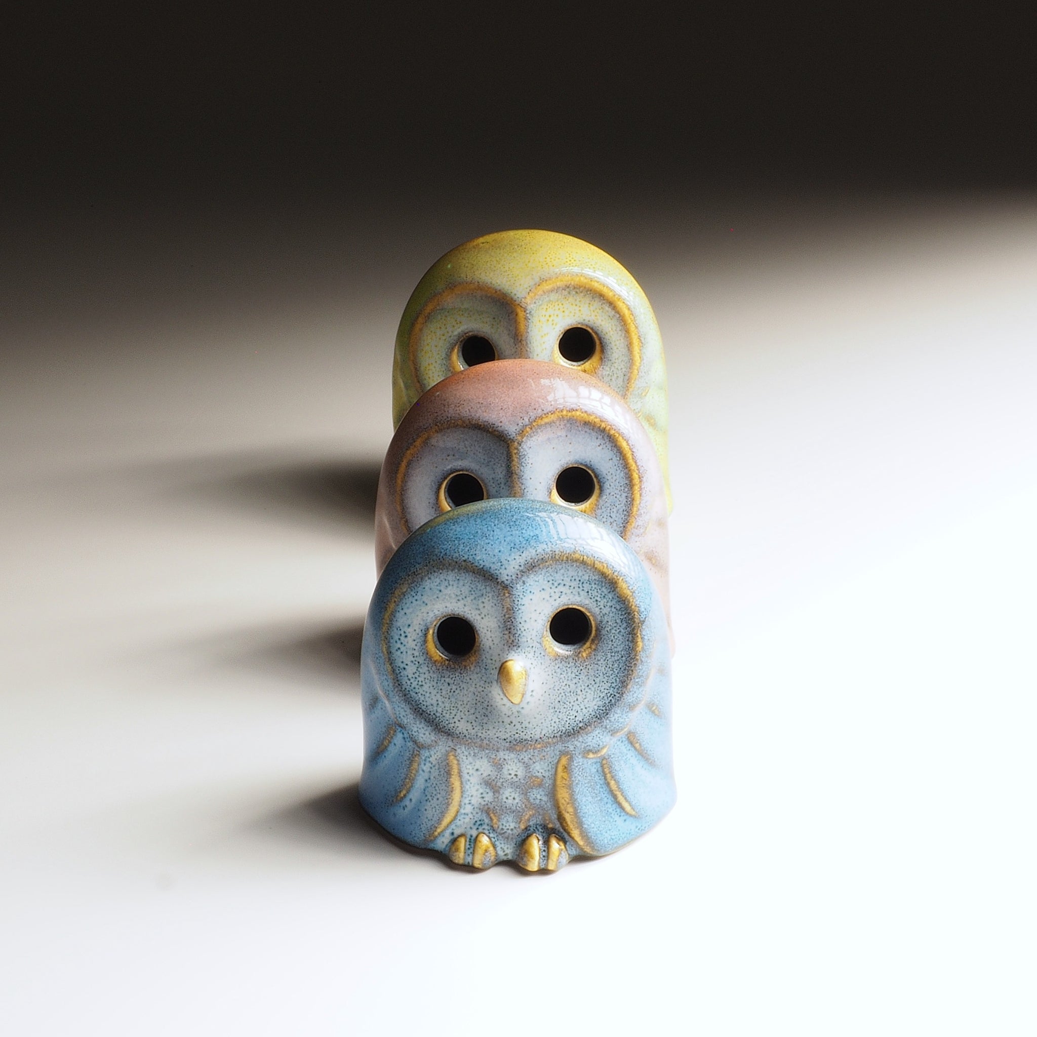 Set of three Engifuku owl figurines by Kazuhiro Kojima stacked in a row: blue, pink, and yellow speckled glazes, hollow eyes.