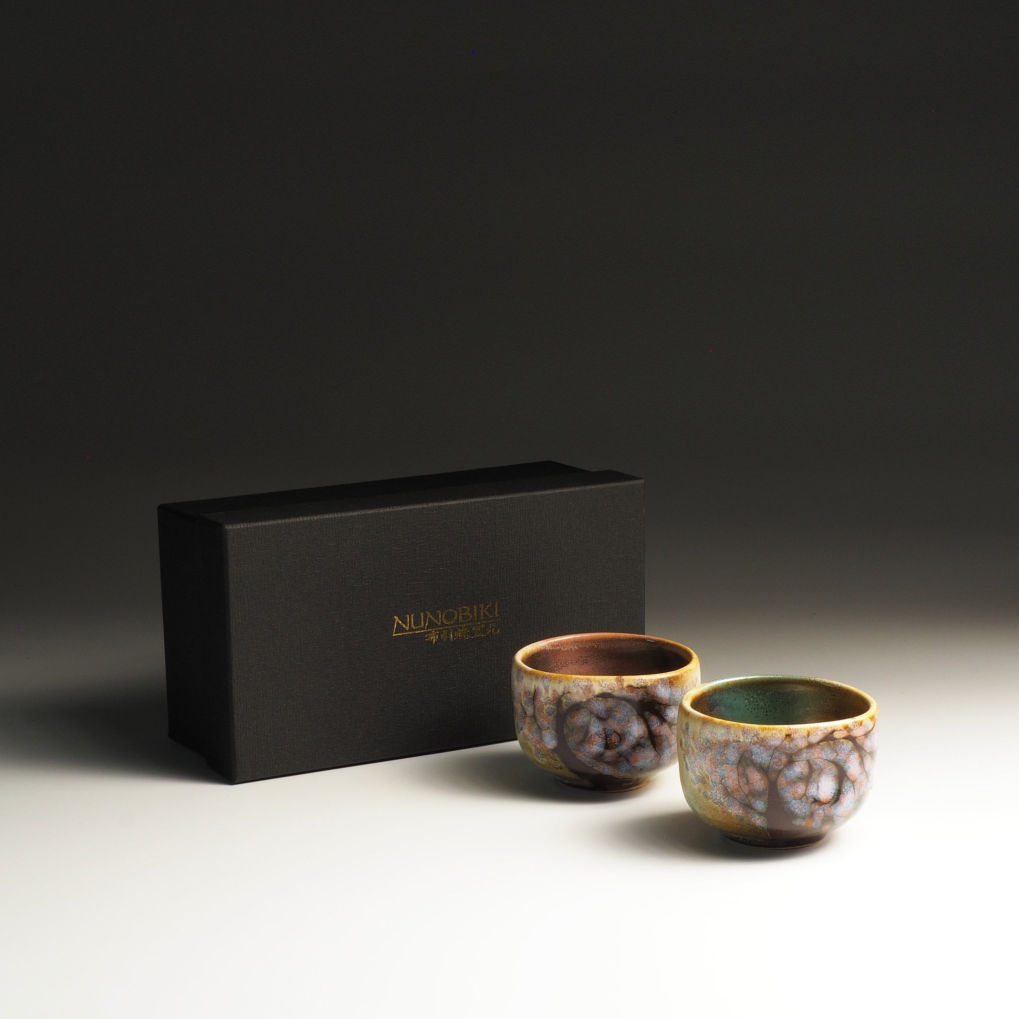 Two Japanese pottery sake cups with a black box on a gradient background