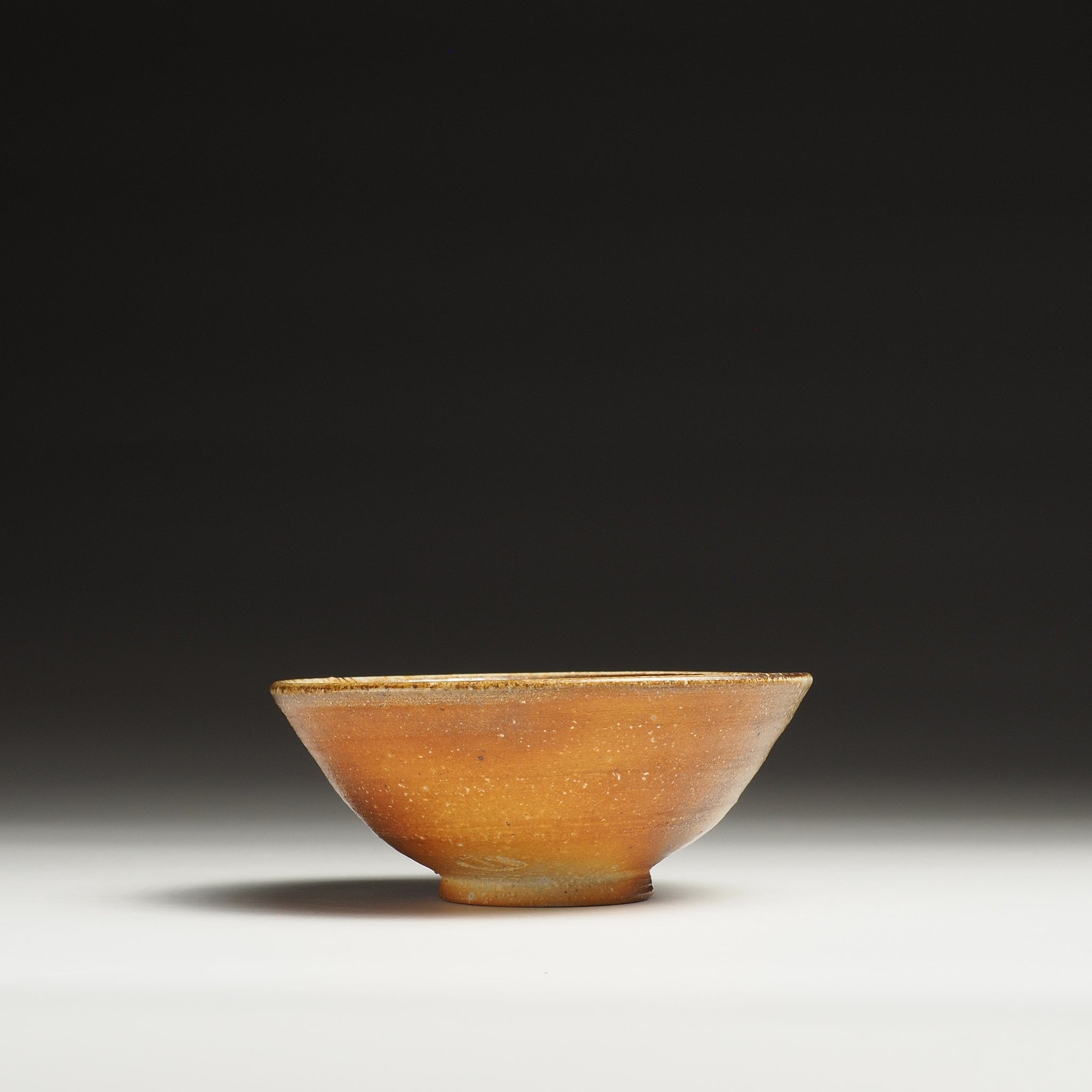 Side view of a Mizuho handmade Shigaraki rice bowl showing its tapered foot and warm amber surface.