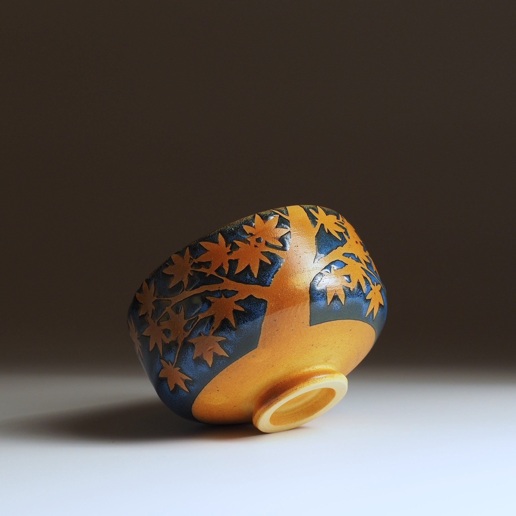 Sōshū matcha bowl tilted forward, momiji tree in negative space wrapping the body, unglazed amber foot ring