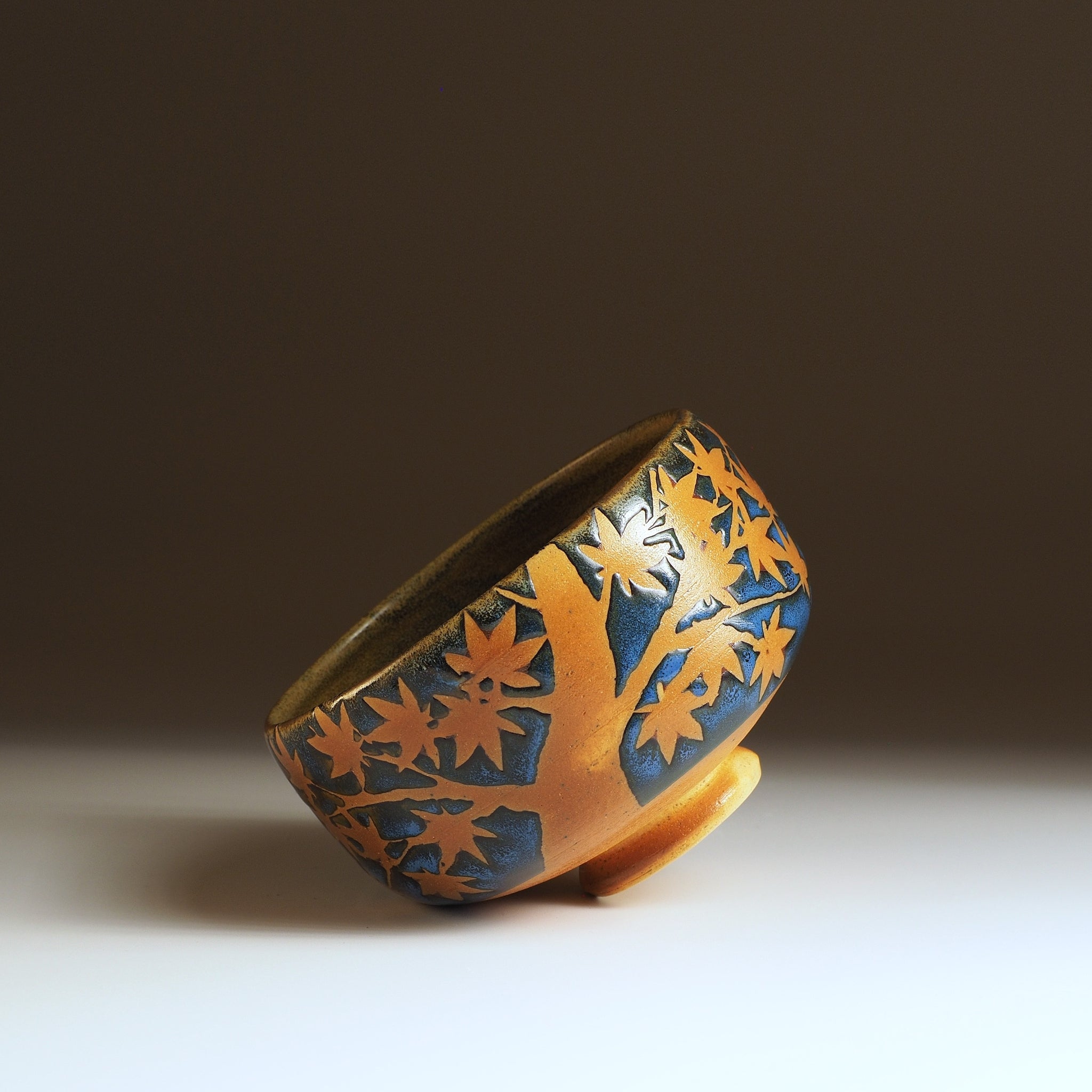 Sōshū matcha bowl tilted, full momiji tree and leaves in negative space, amber clay trunk rising from foot ring
