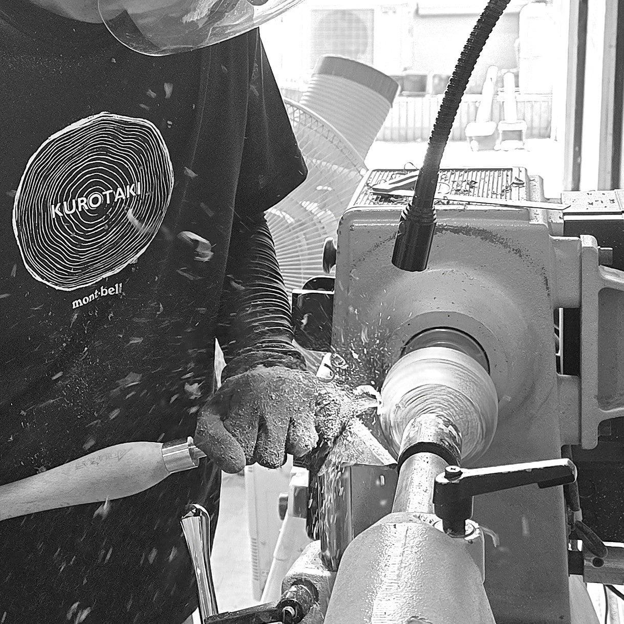 A black and white photo of a woodturner operating a lathe machine, wearing a protective face shield and a black t-shirt. The woodworker holds a chisel to shave off material from a spinning wood blank, with metal shavings flying in the air.